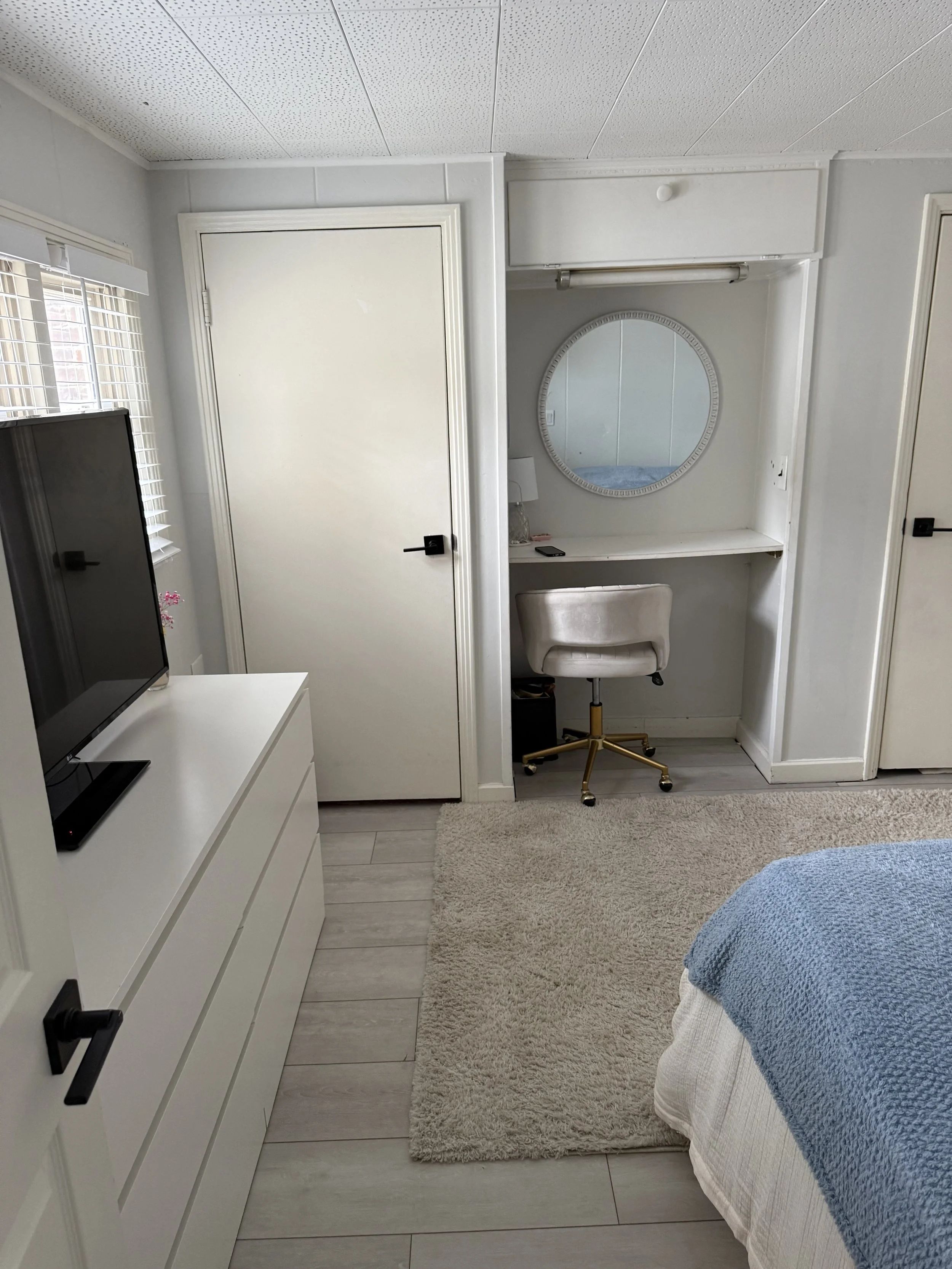 A bedroom with white walls, light wood flooring, and a window with blinds. There is a white dresser with a television on top, a small beige shag carpet, and part of a bed with a blue blanket. A built-in desk area with a round mirror, a lamp, a chair,