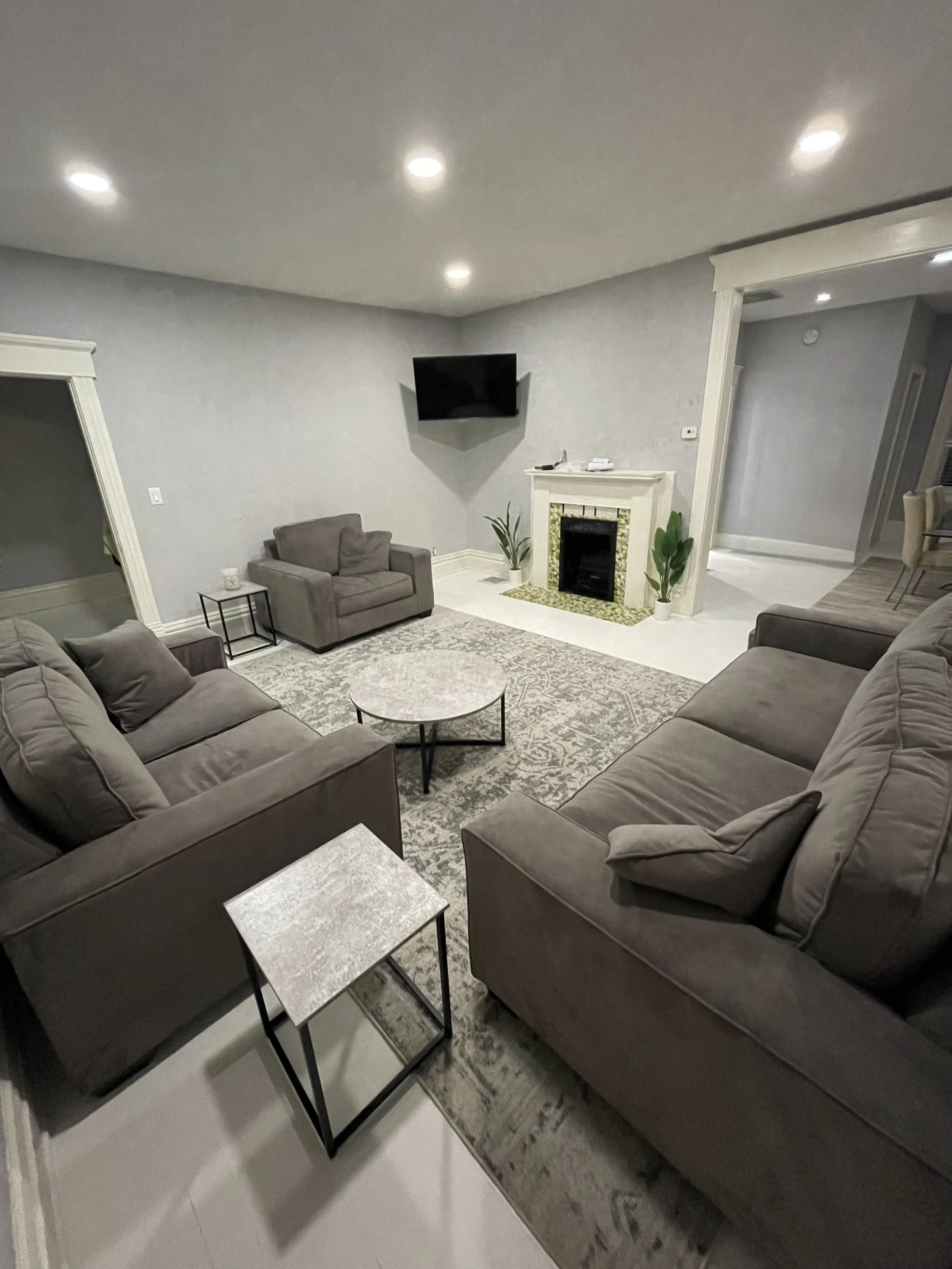 Living room with beige couches, a marble-top side table, a carpet, a wall-mounted TV, a fireplace, two potted plants, and recessed ceiling lights.
