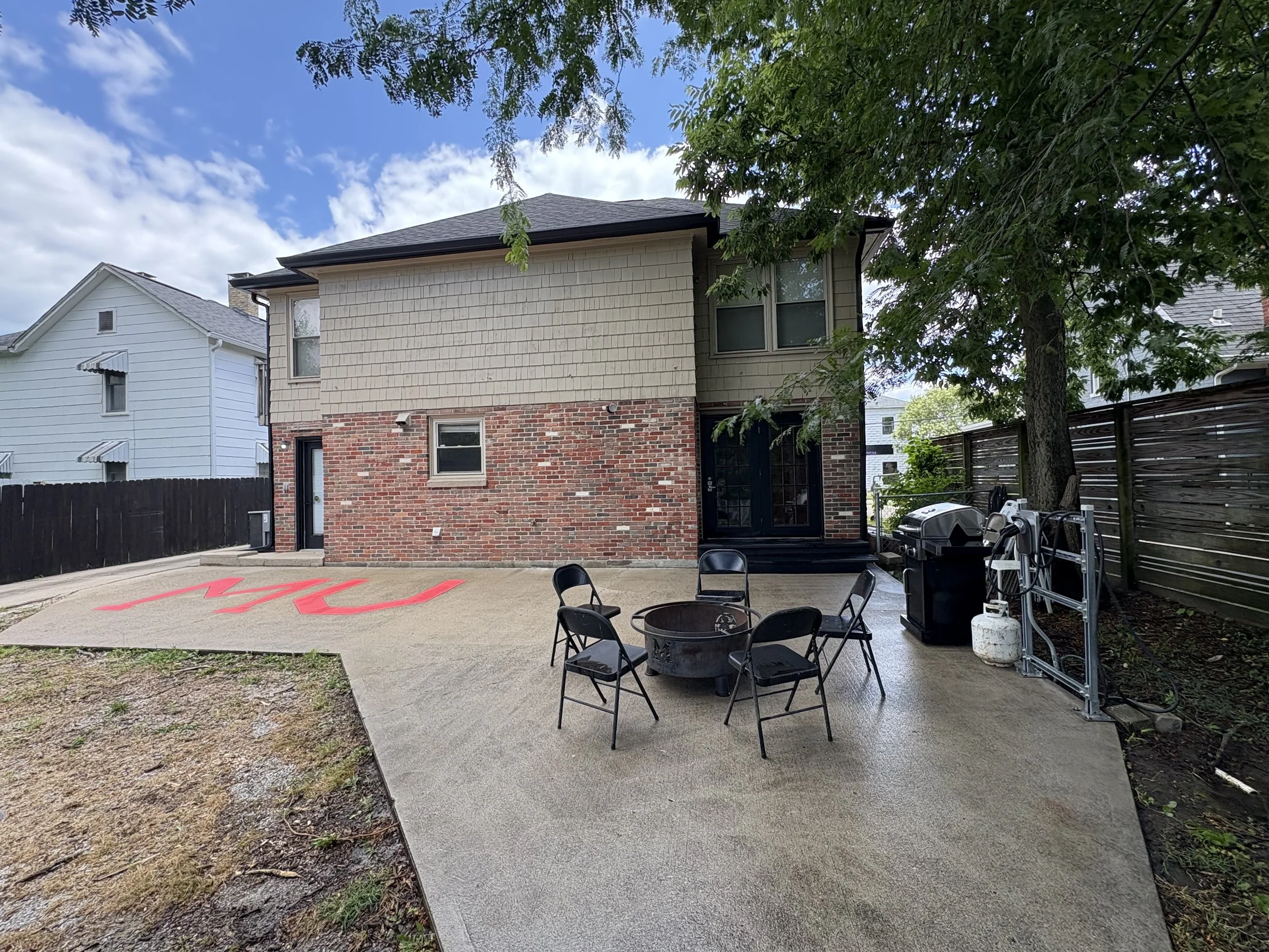 The backyard of a house with a concrete patio, black chairs, and a grill. There is a large tree with green leaves, a wooden fence, and neighboring houses visible in the background. The sky is partly cloudy.