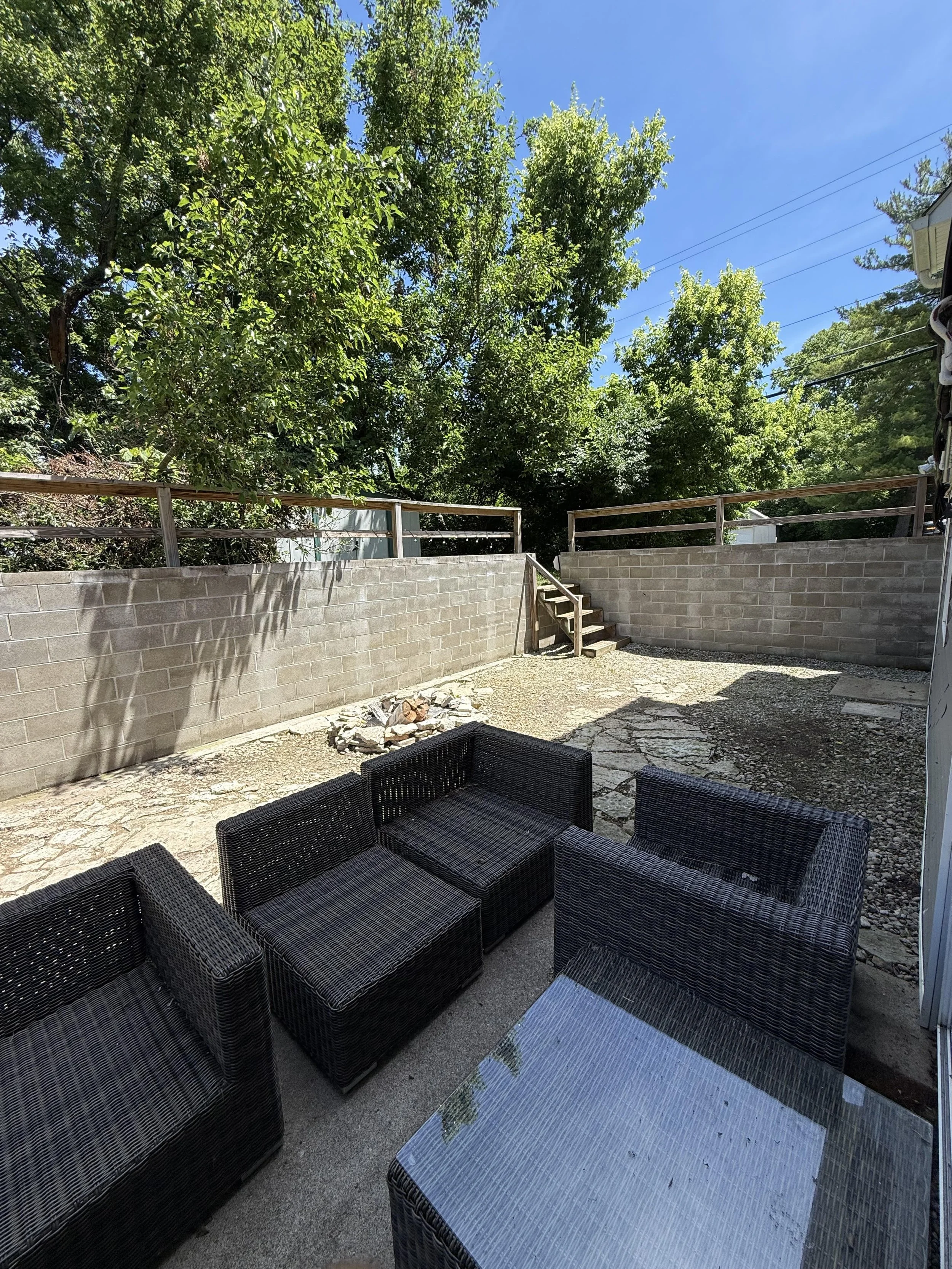 A backyard patio with outdoor black wicker furniture including sofas and a glass-top table, surrounded by a concrete block wall, with stairs leading to a higher level, trees and a clear blue sky in the background.