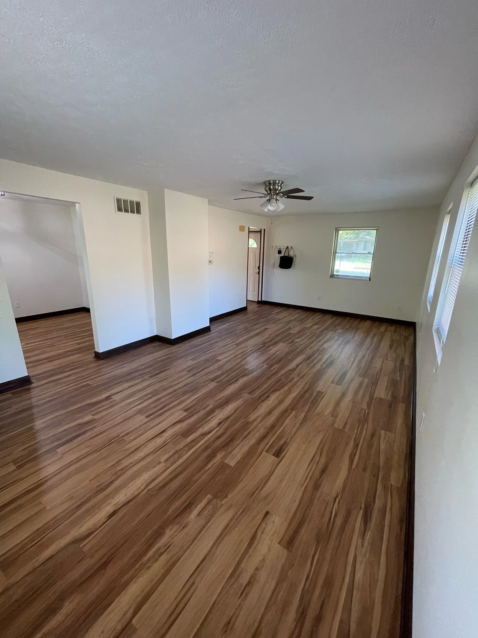 Empty living room with wood laminate flooring, white walls, a ceiling fan, and windows with blinds.