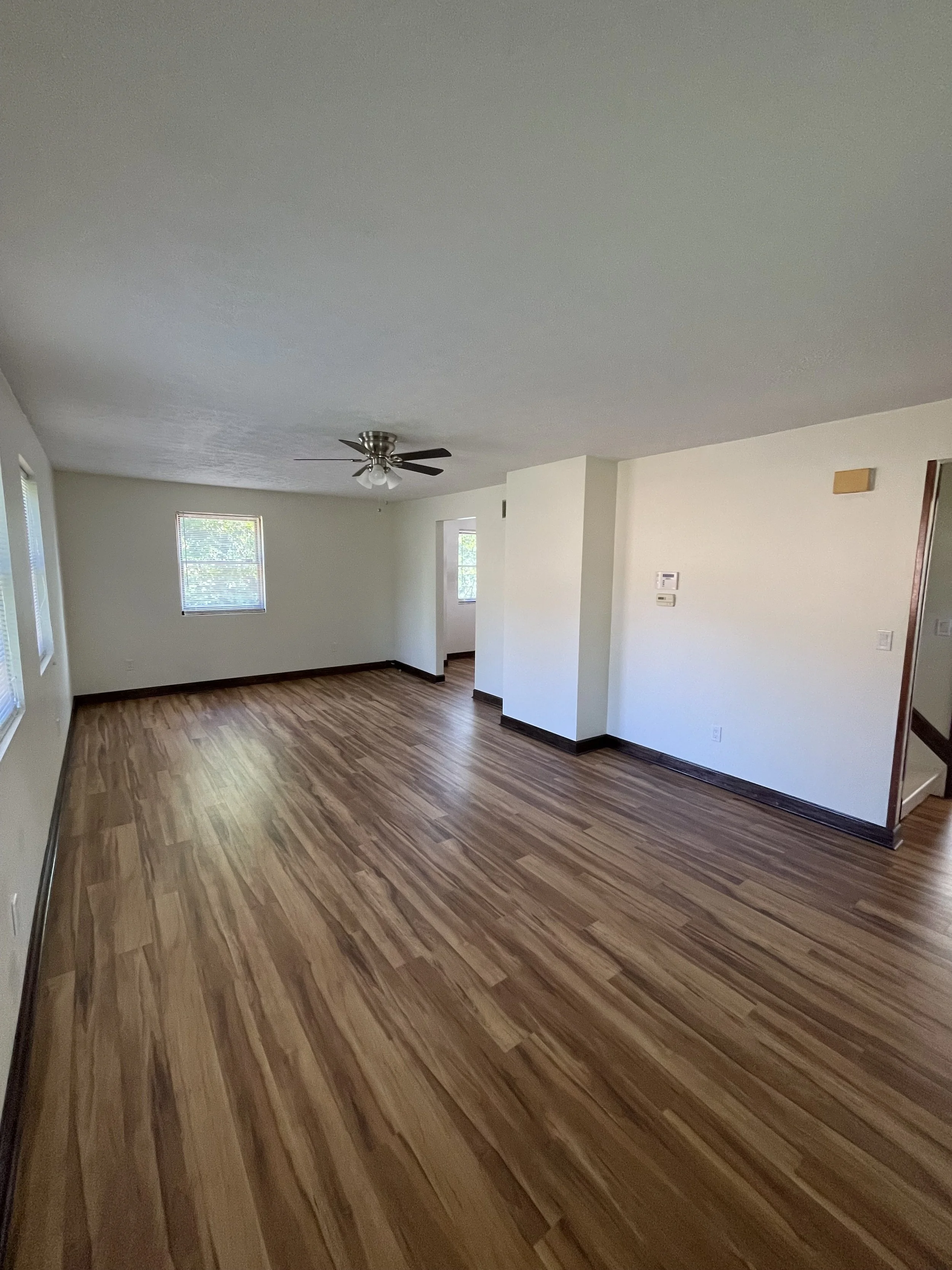 Empty living room with hardwood floors, white walls, windows with blinds, and a ceiling fan with light.