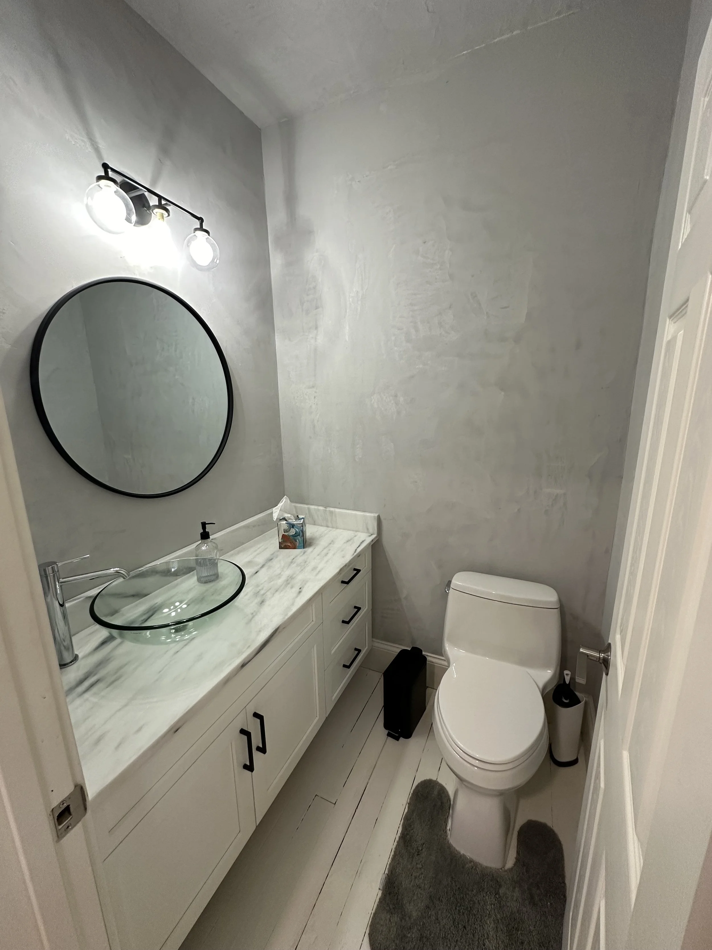 A small modern bathroom with a white vanity, marble countertop, and a round mirror. There is a glass vessel sink and a black framed mirror above the sink. Lighting fixtures are on the wall, and a toilet with a gray rug in front is visible. Toilet pap
