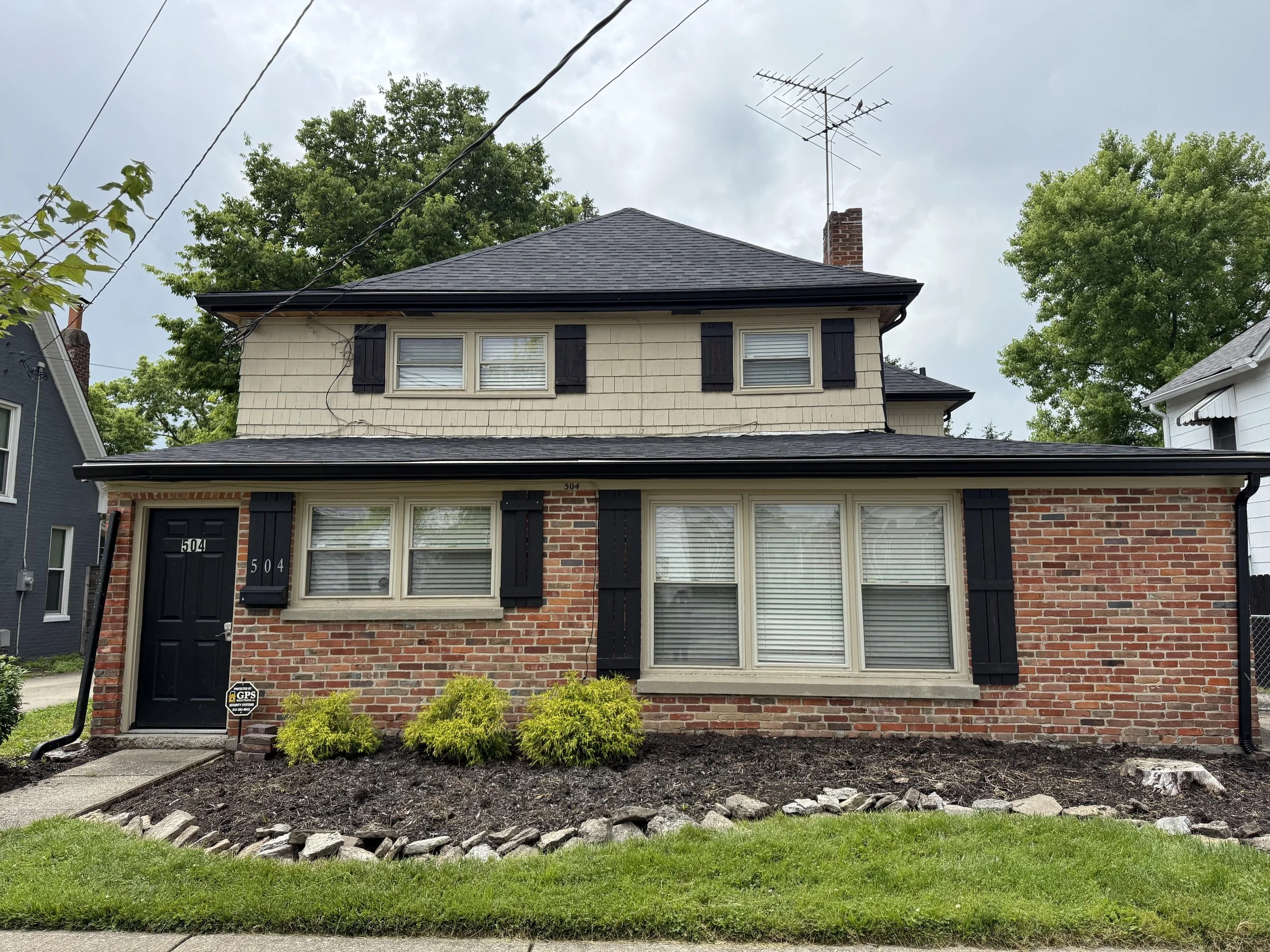 A two-story house with cream-colored siding, black shutters, and a brick foundation, with multiple windows and a front door. The house has a gray shingled roof with a chimney, surrounded by green trees and a small landscaped garden with bushes and mu
