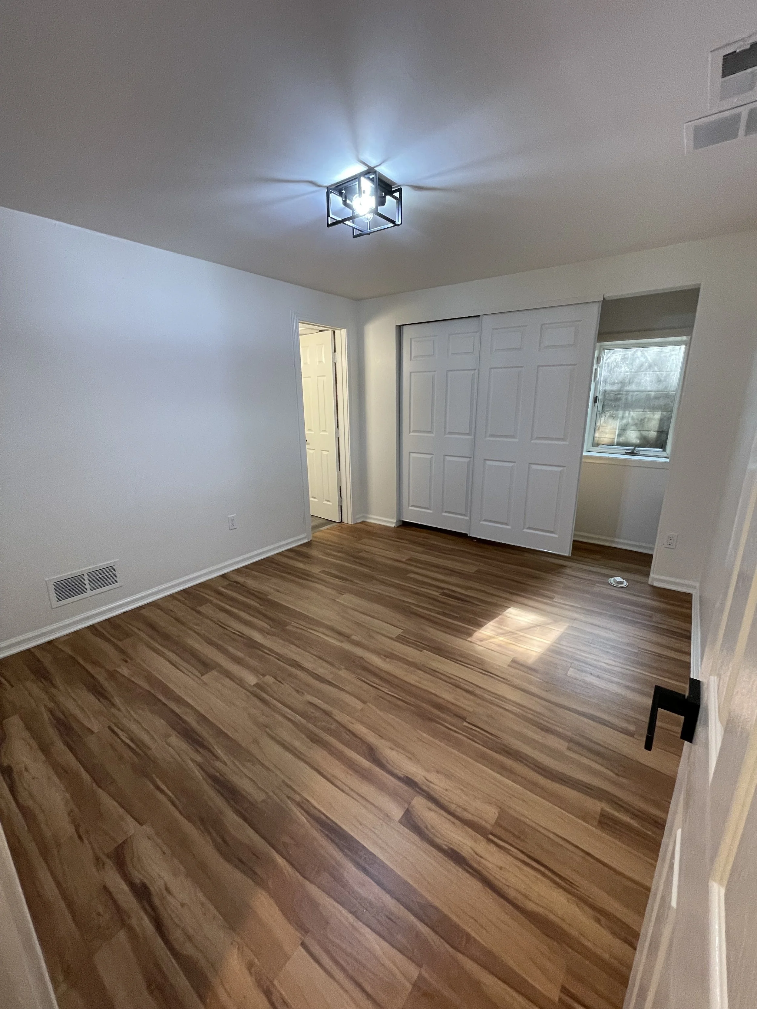 A room with light-colored walls, a ceiling light fixture, a window, a closet with doors, and wood flooring.