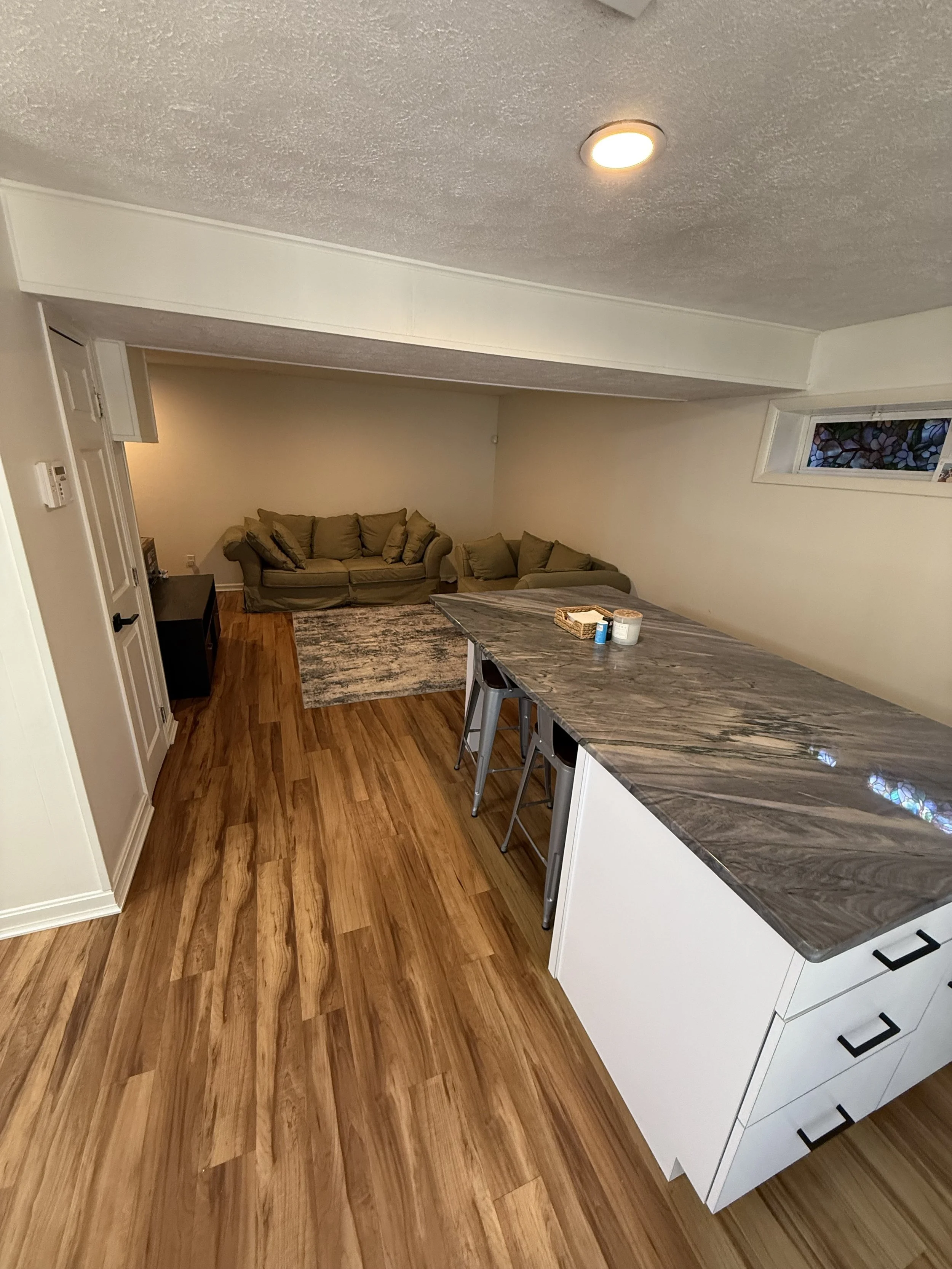 A living room and kitchen area with hardwood floors, a sofa with multiple cushions, a small black table, a window with stained glass, a granite kitchen island with some items on it, and a white cabinet with black handles.