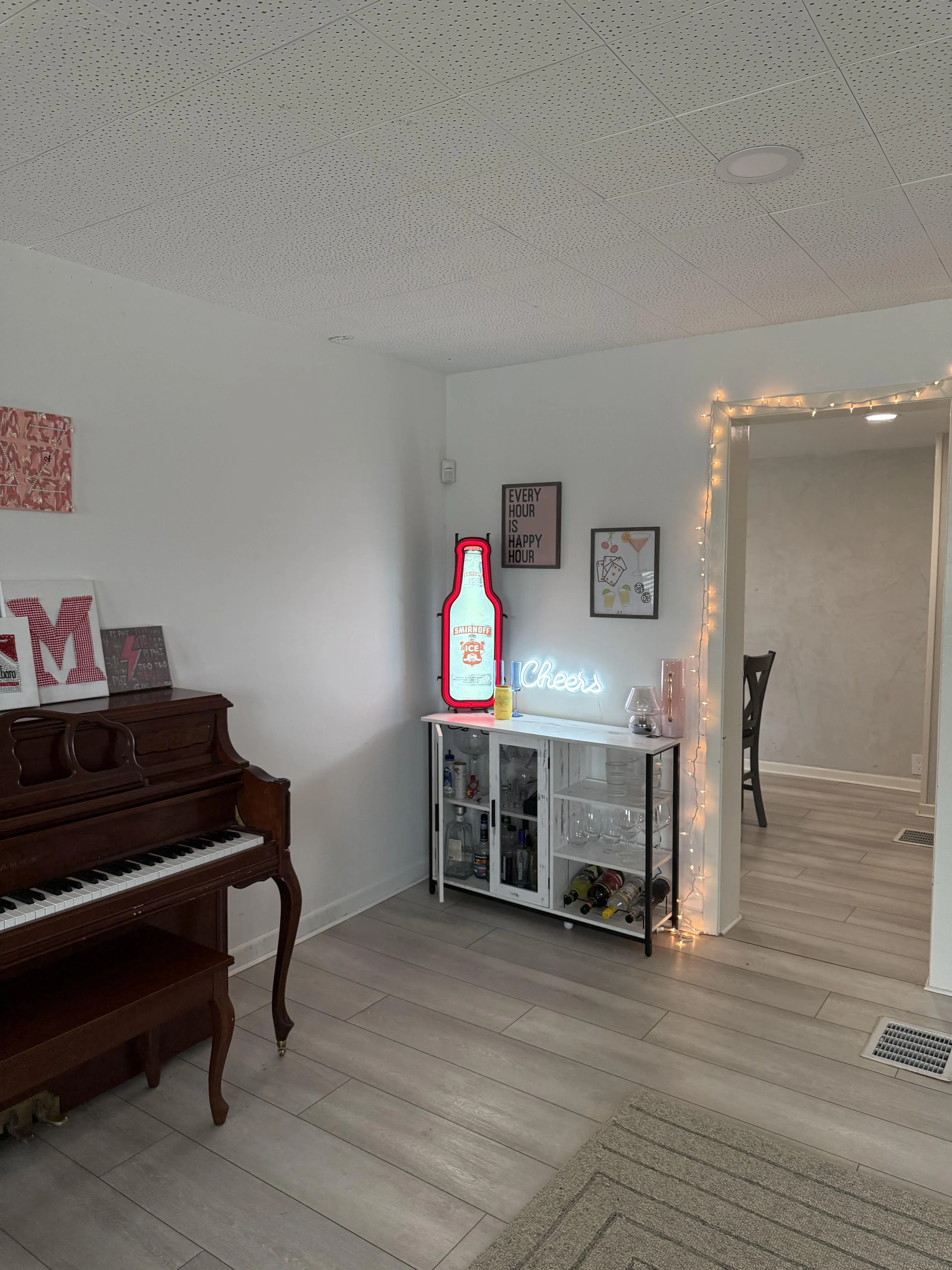 Interior of a house with a piano on the left, a bar cart with bottles and glassware on the right, framed artwork on the wall, and string lights around a doorway.