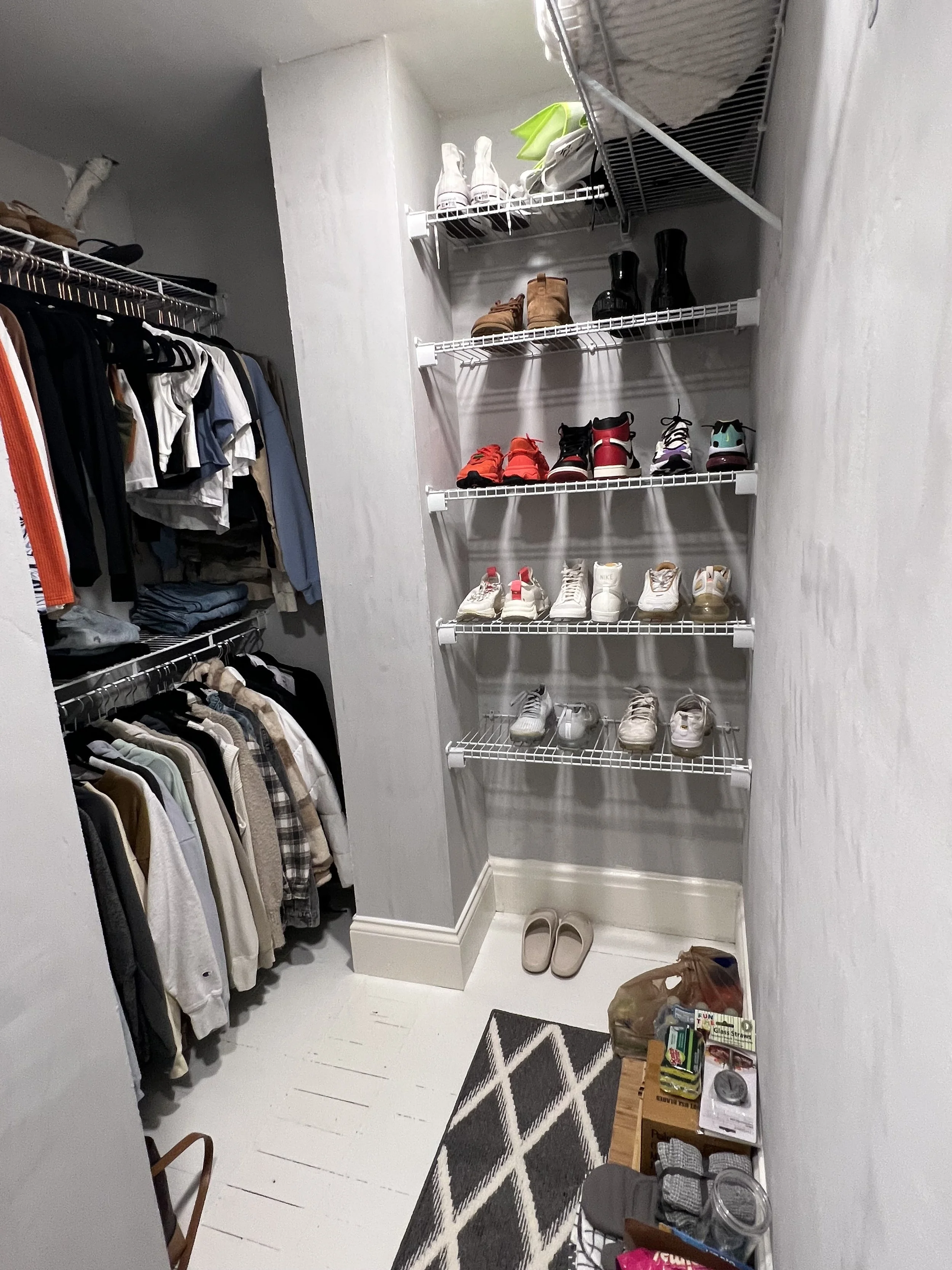 A walk-in closet with shelves and racks holding shoes, clothing, and accessories, near a small table with items on it and a patterned rug on the floor.