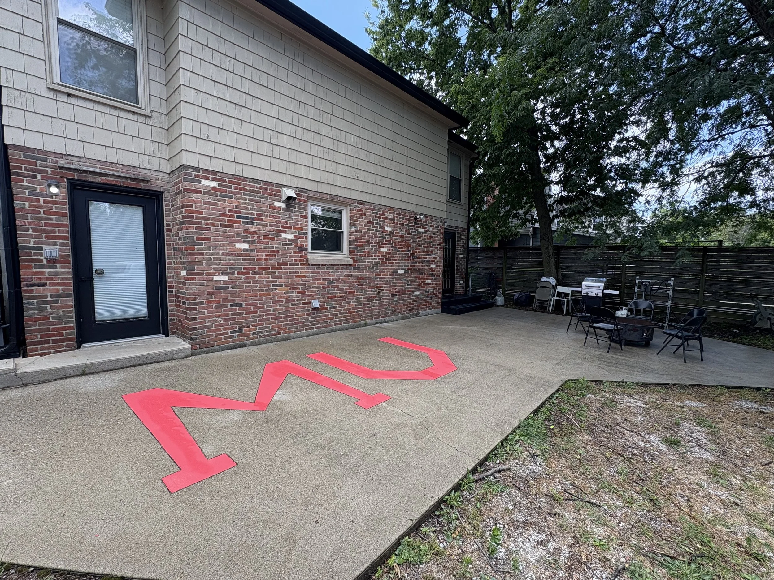 Back patio area with a concrete surface, neighbor's brick and siding house wall, a door with window blinds, outdoor seating including chairs and a grill, large trees providing shade, and a pink arrow marking the ground.