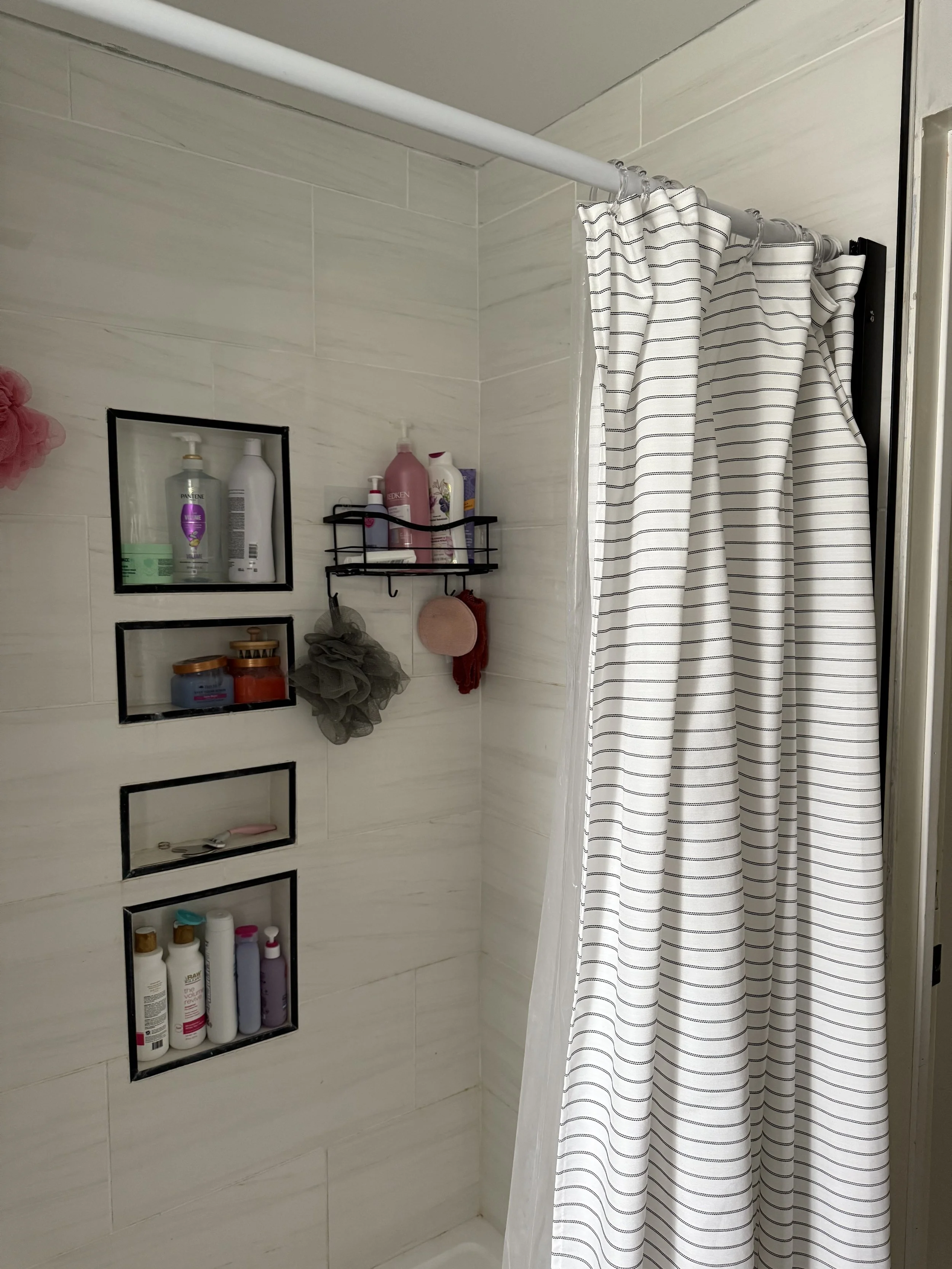 Inside a shower with a white and black striped shower curtain. Recessed shelves with toiletries such as bottles of shampoo, conditioner, and body wash.