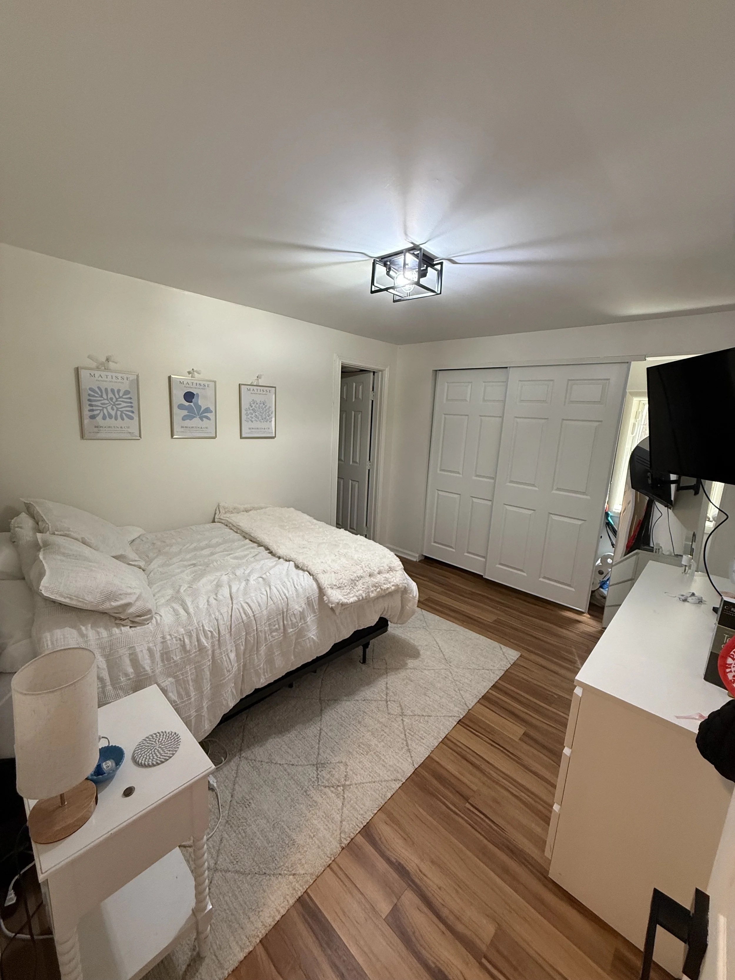 A bedroom with a bed covered in white bedding and pillows, a nightstand with a lamp and decorative items, framed artwork on the wall, a closet with sliding doors, a flat-screen TV mounted on the wall, and wooden flooring.