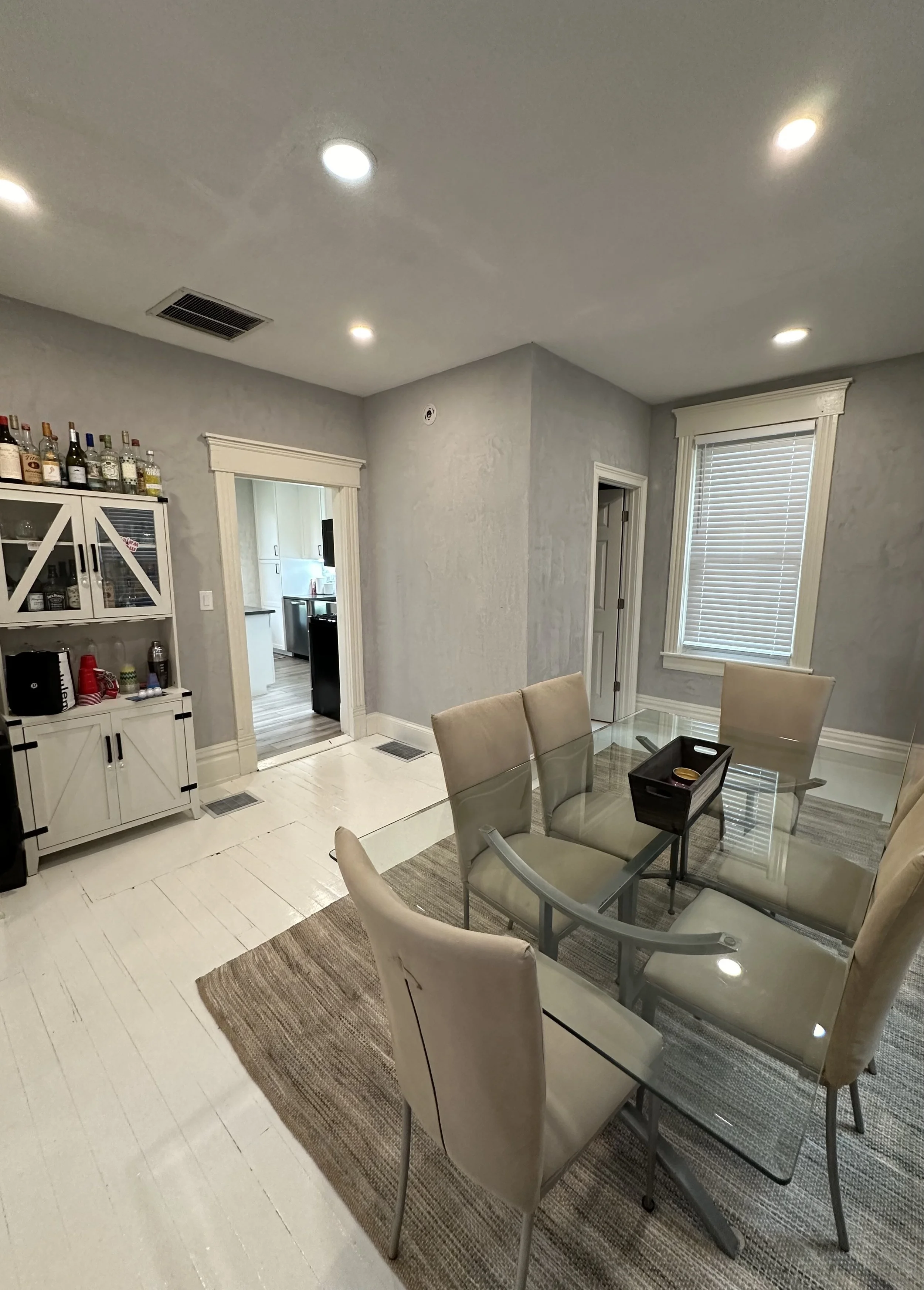 A dining area with a glass table and beige chairs, a window with blinds, a white cabinet with liquor bottles, and a view into a kitchen.