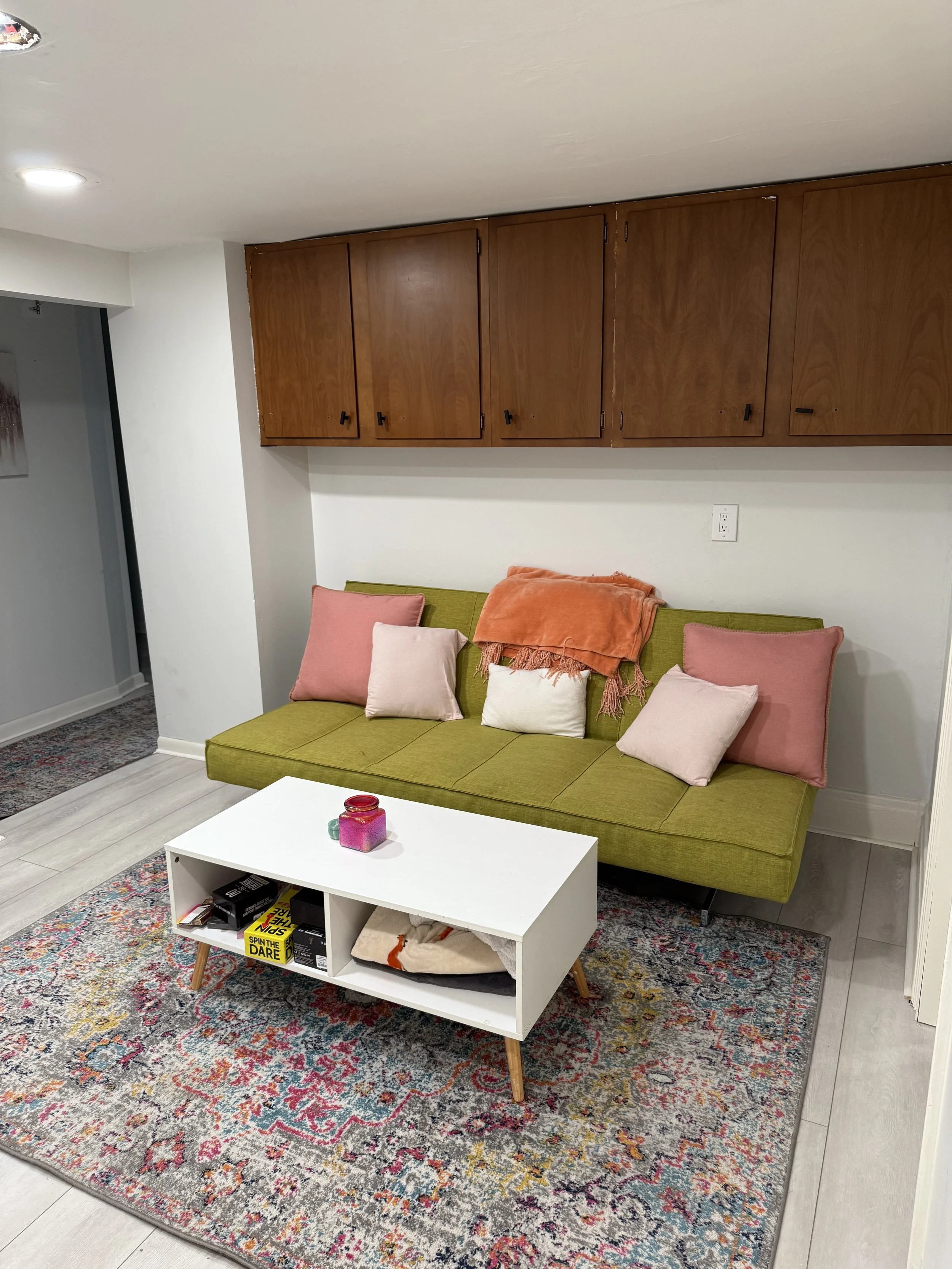 Living room with a green sofa, pink and white accent pillows, wooden cabinets on the wall, a white coffee table with decorative items and books, a colorful patterned rug, and part of a doorway to another room.