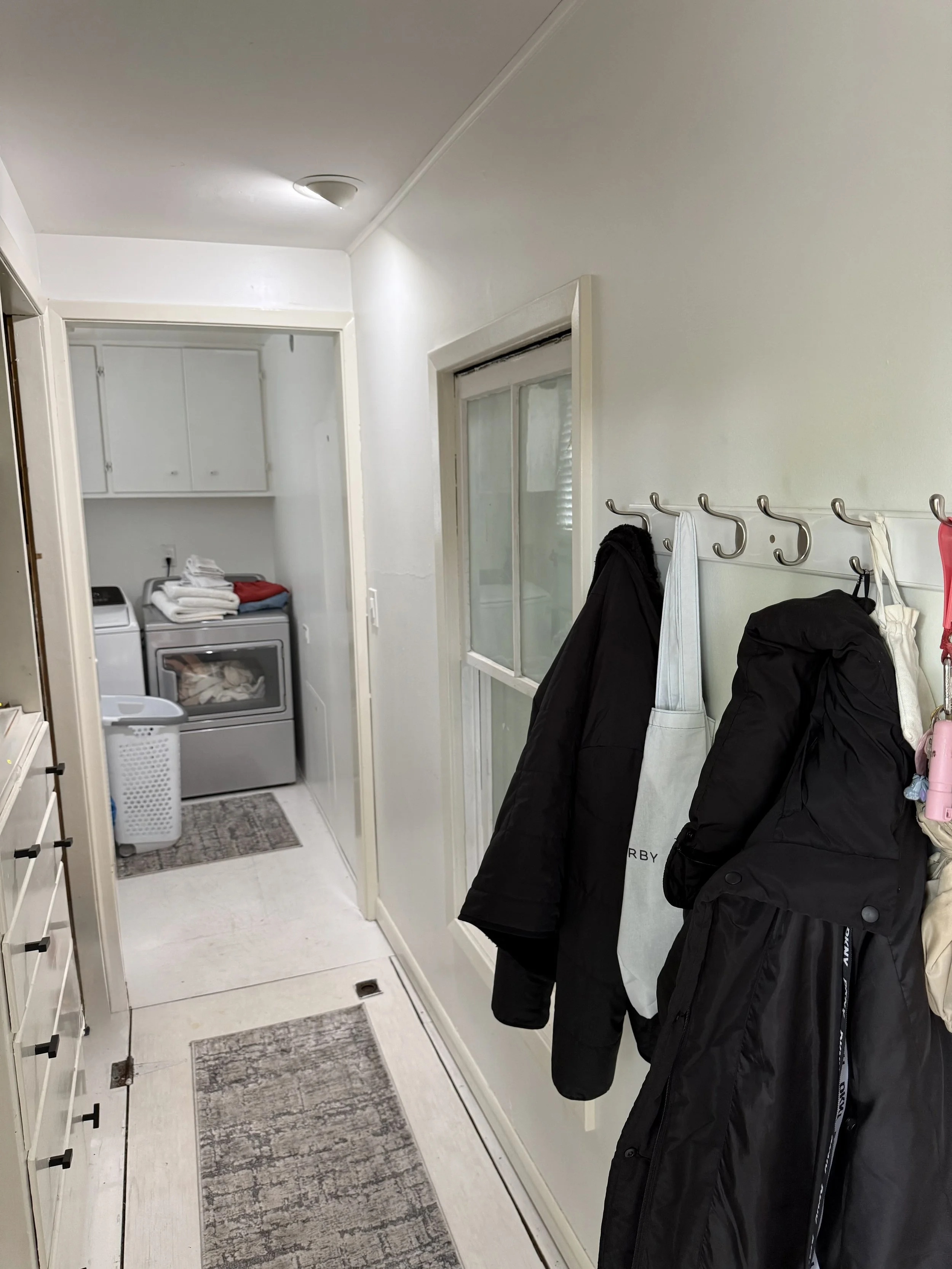A laundry room with a washer and dryer, some folded clothes on top of the washer, and a laundry basket. A coat rack with hanging jackets and bags is on the right side. A door with a window leads to the outside. The room has white walls and tiled floo
