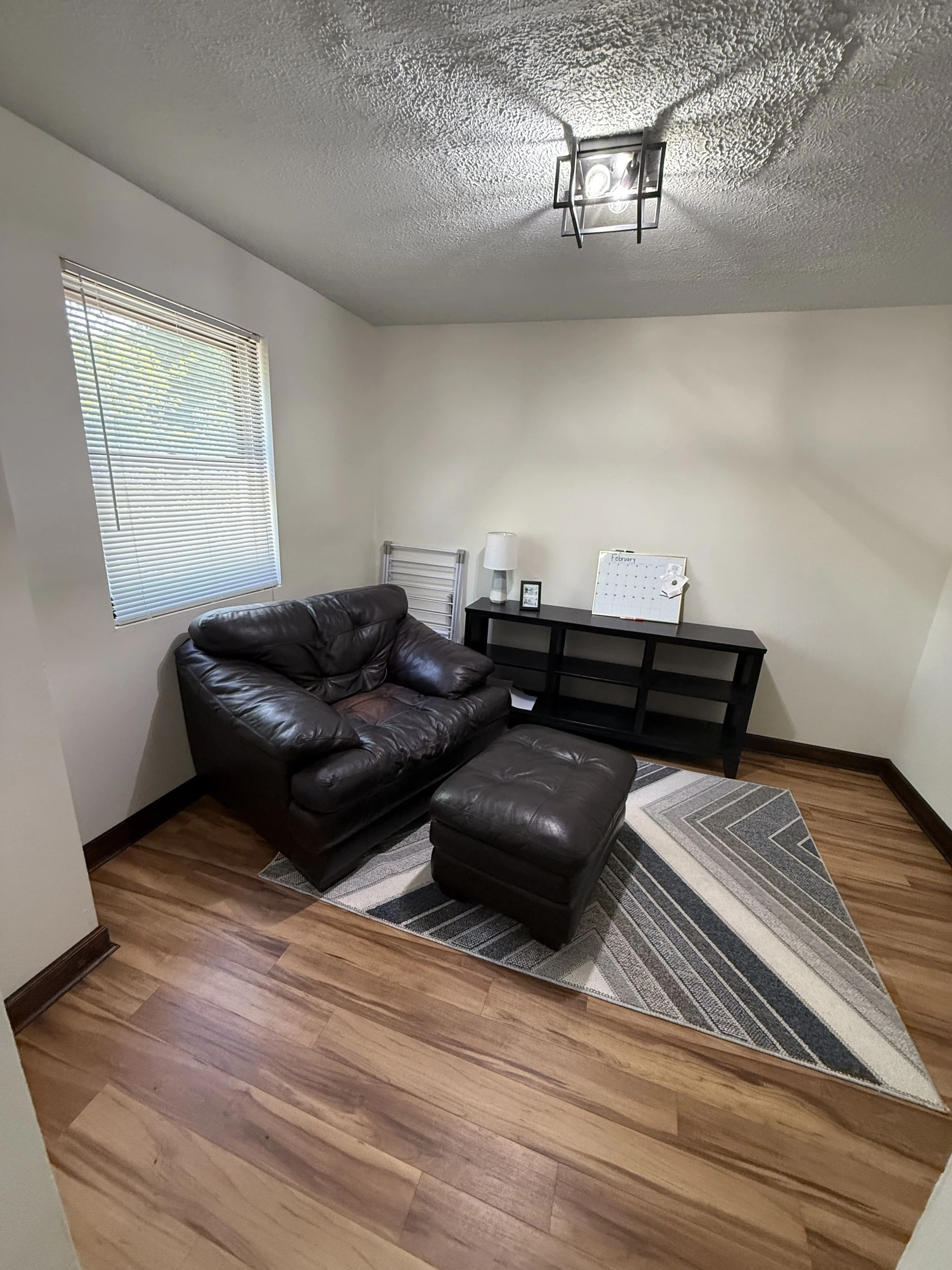 A living room with a brown leather sofa and matching ottoman, a black side table with a white lamp, a small framed photo, a whiteboard calendar, a window with blinds, a patterned area rug, and wooden flooring.