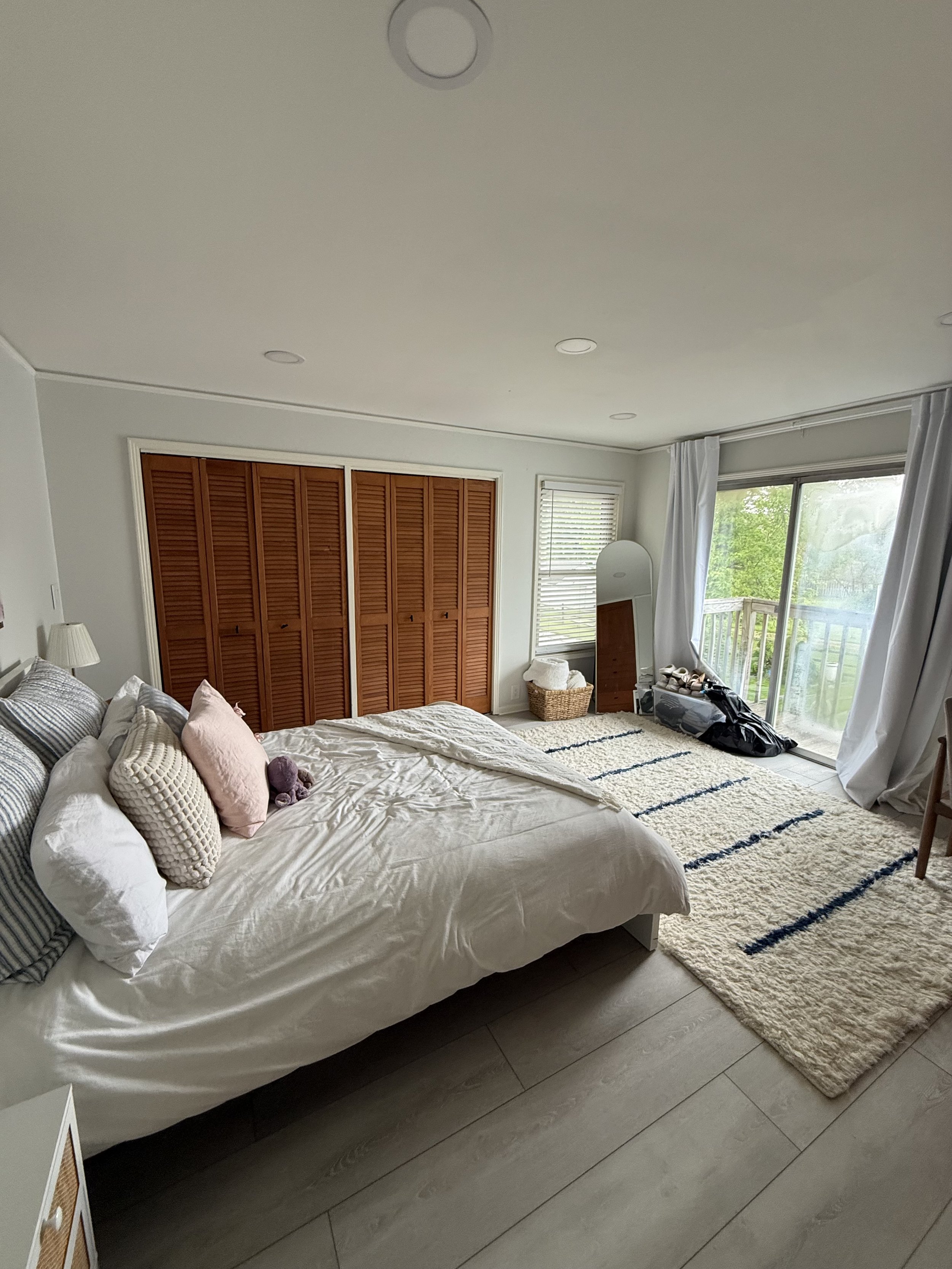A bedroom with a bed, pillows, a rug, and a sliding glass door leading to an outdoor area with greenery. There is a closet with louvered wooden doors, and a mirror next to the window. Items such as shoes and bags are on the floor near the door.