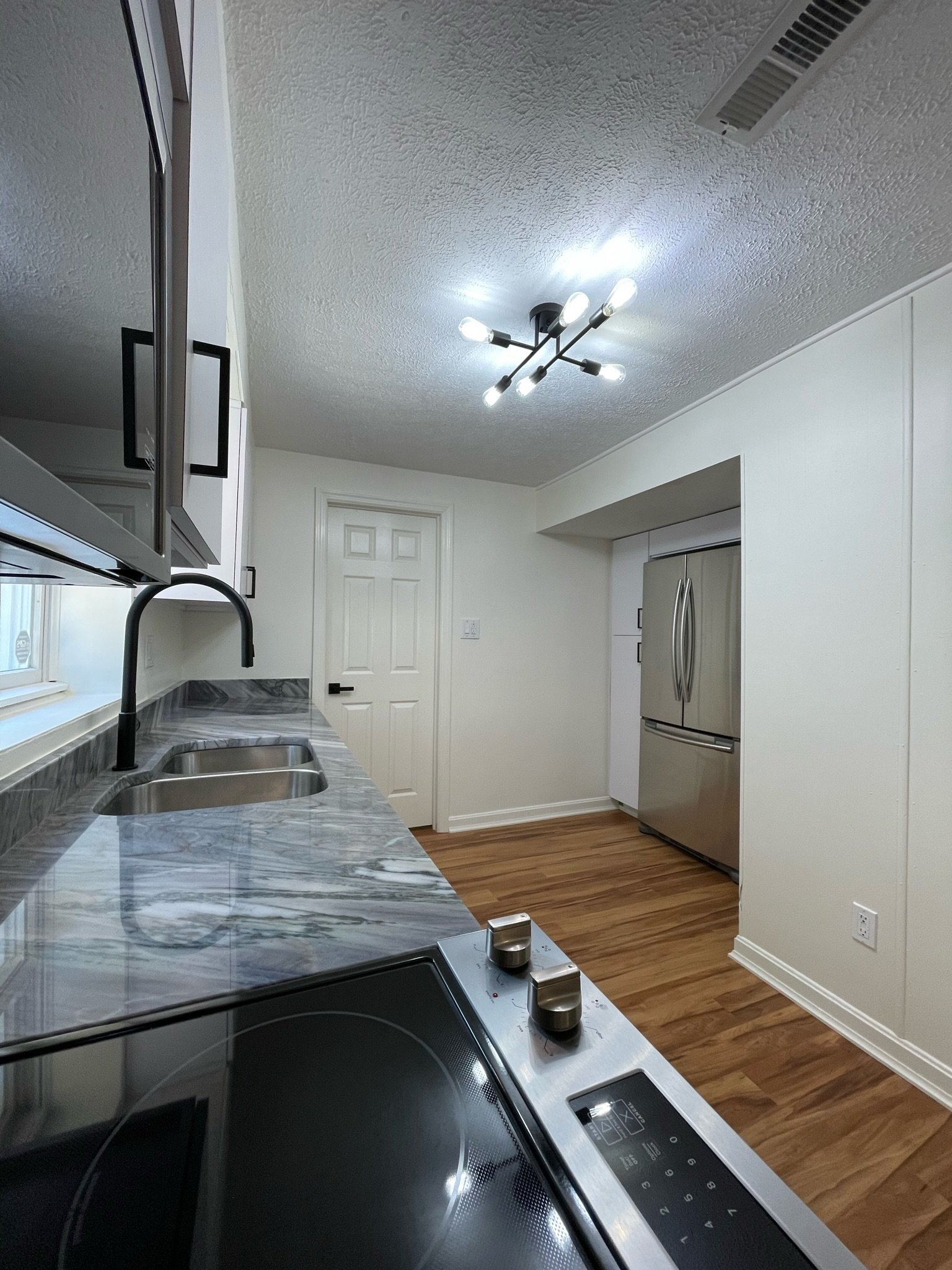 Modern kitchen with a marble countertop, stainless steel refrigerator, electric stove, black sink faucet, light-colored walls, wood flooring, overhead light fixture, window, and white door.