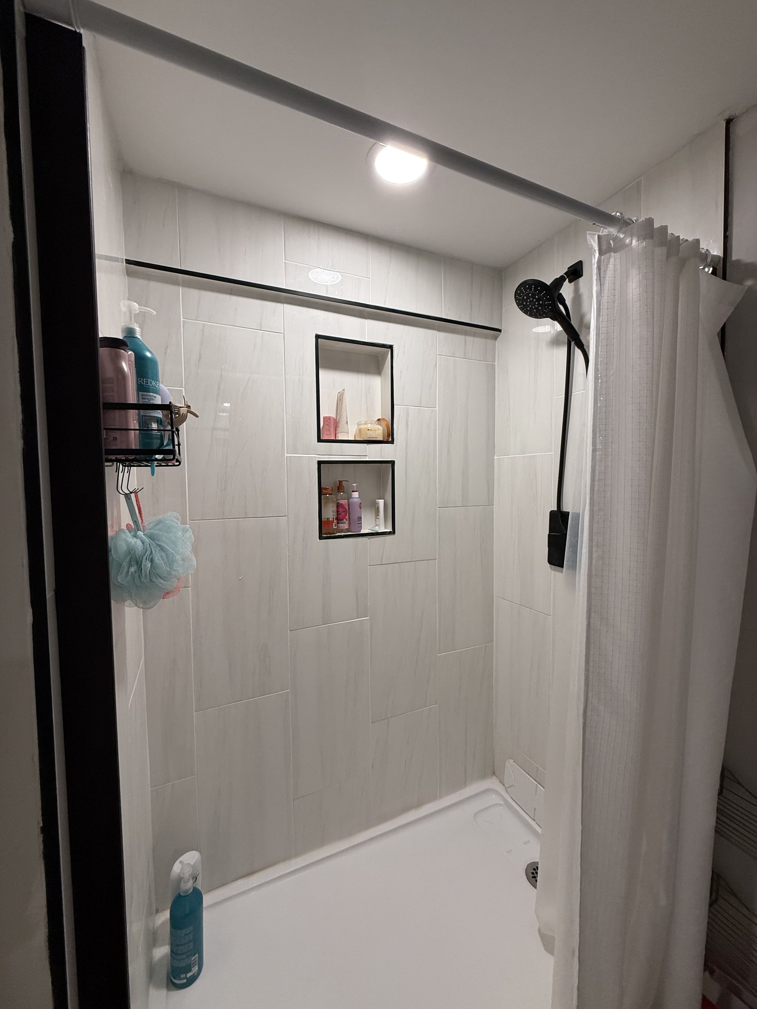 A small shower stall with white tiled walls, a black shower head, and a corner shelf holding various toiletries. There is a white shower curtain partially drawn, and a bottle of blue shampoo or body wash on the shower floor.
