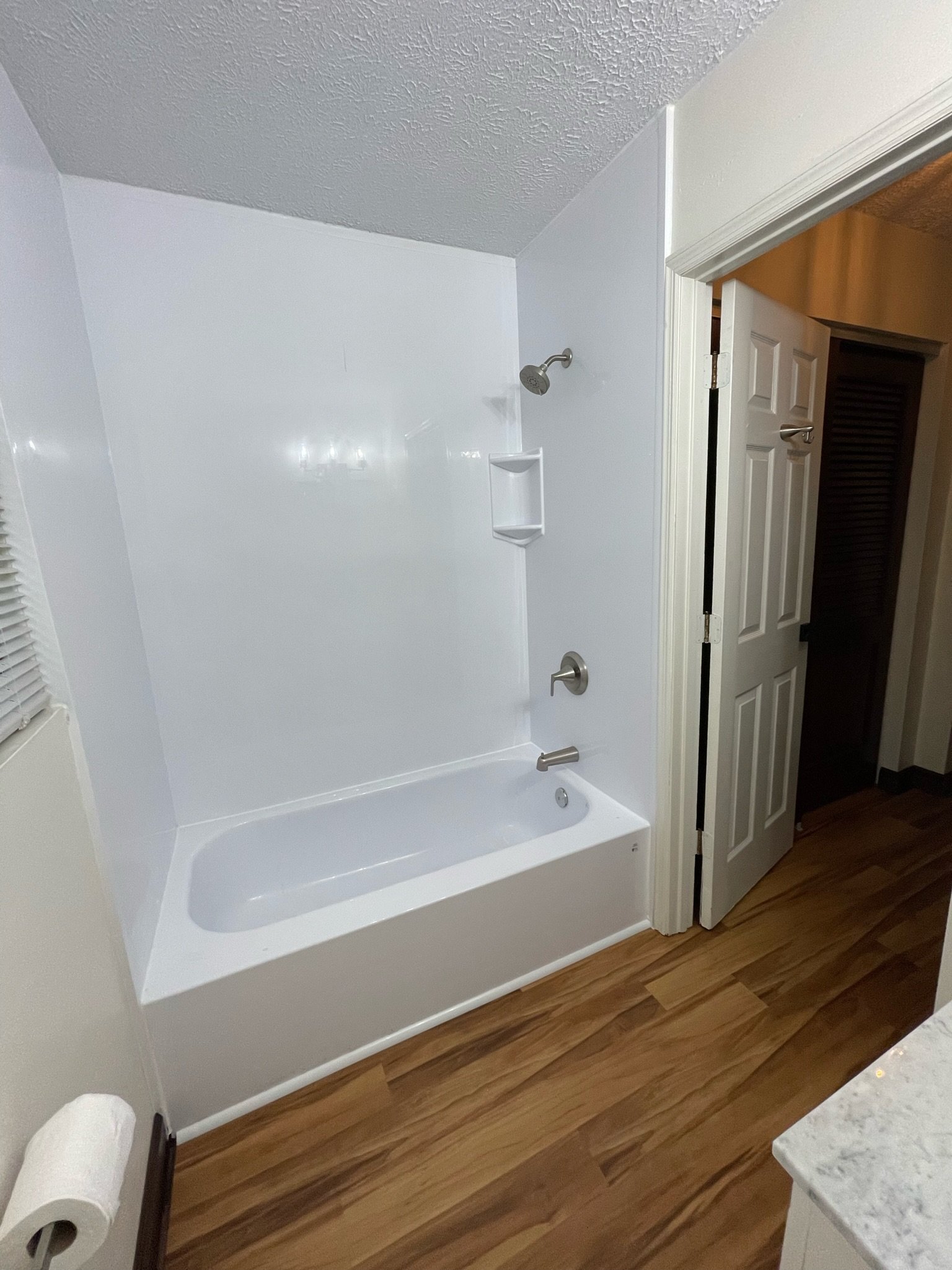 Bathroom with a white bathtub, showerhead, and tile surround. Toilet paper roll on holder, wood flooring, white walls, and a partially open door leading to another room.