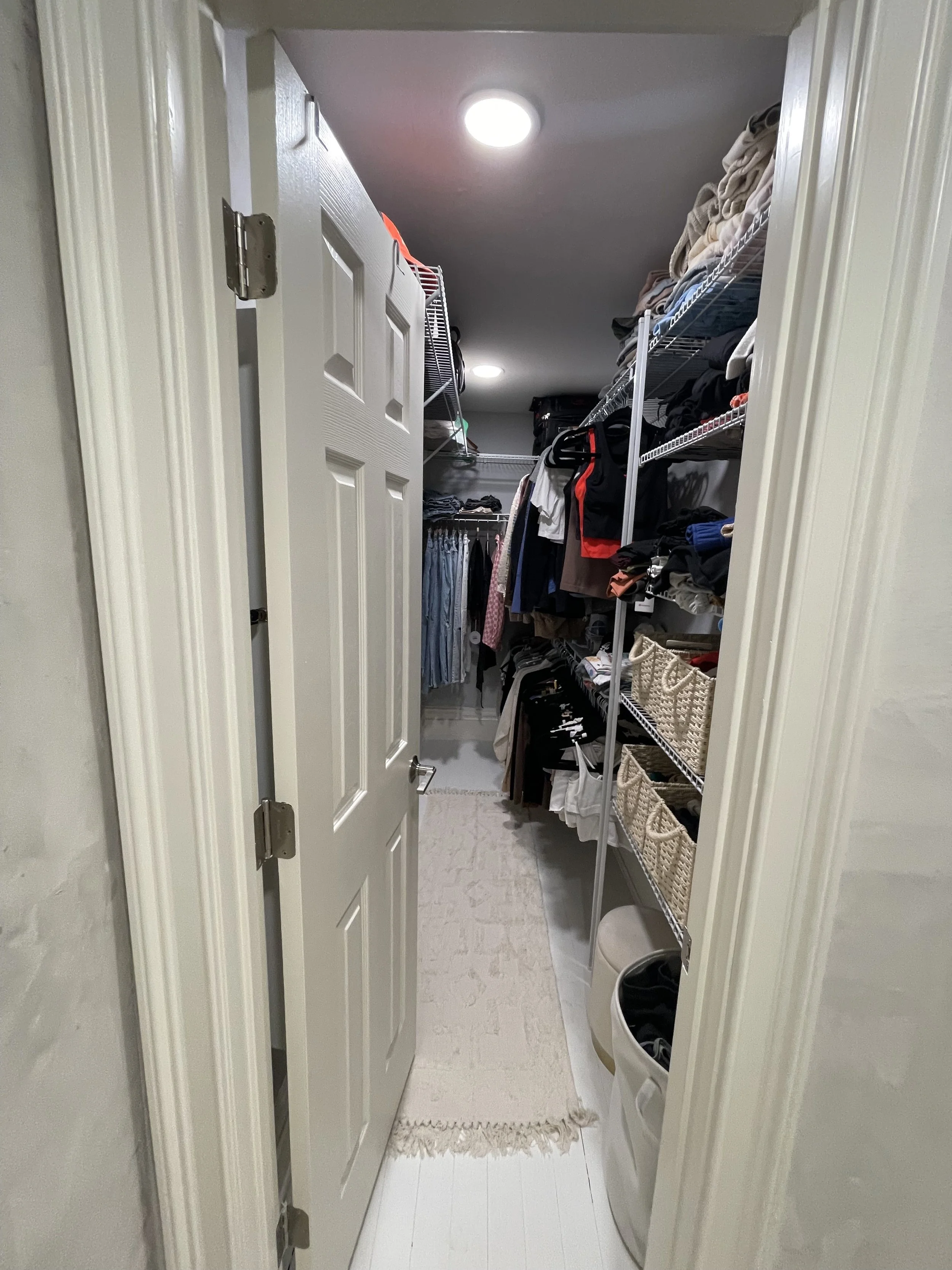 A small walk-in closet with wire shelves holding folded clothes, baskets, and bags, with a laundry basket on the floor and a rug near the entrance.