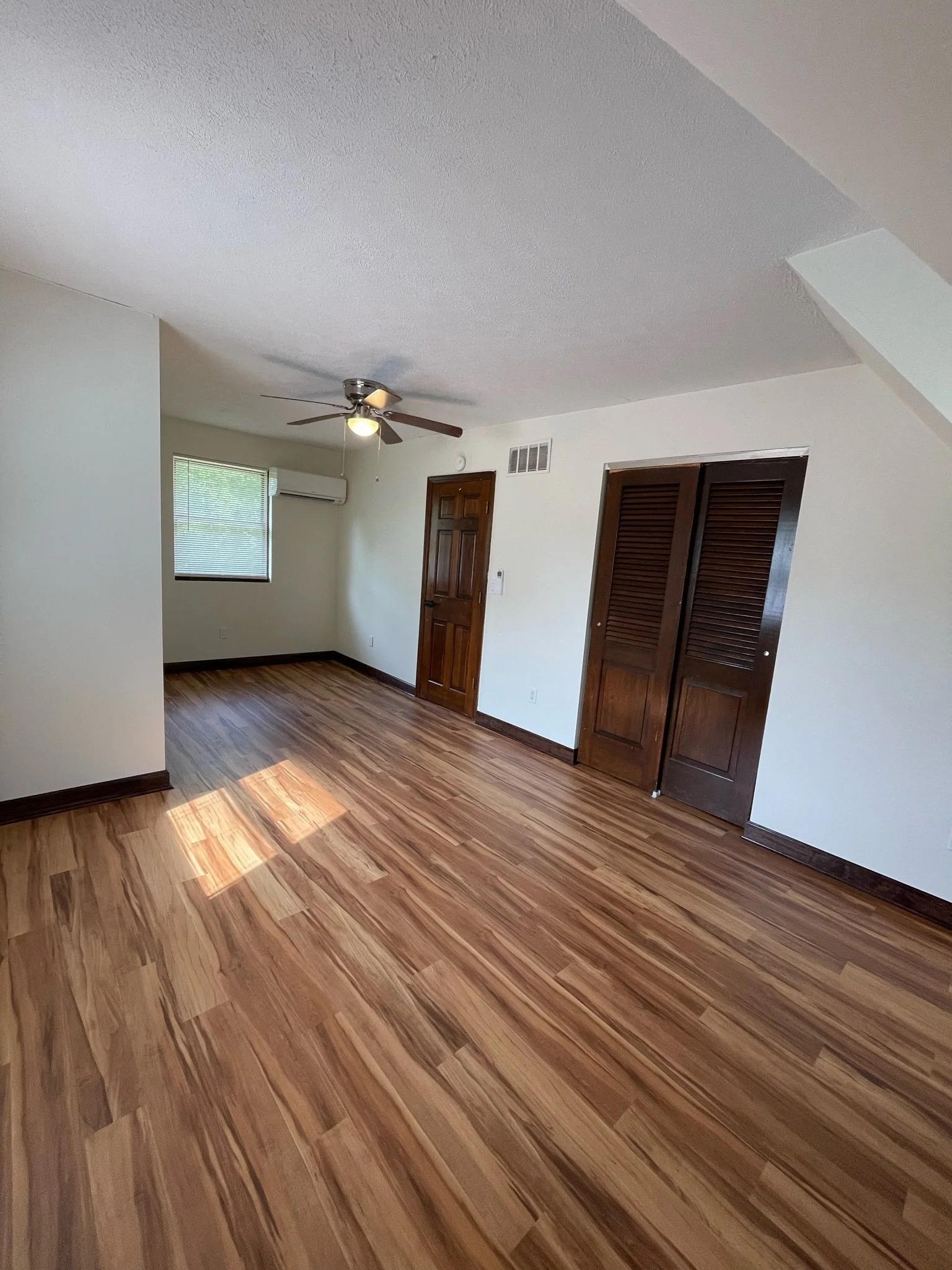 Empty room with wooden floors, white walls, a ceiling fan, a window with blinds, a wooden door, and closet with louvered doors.