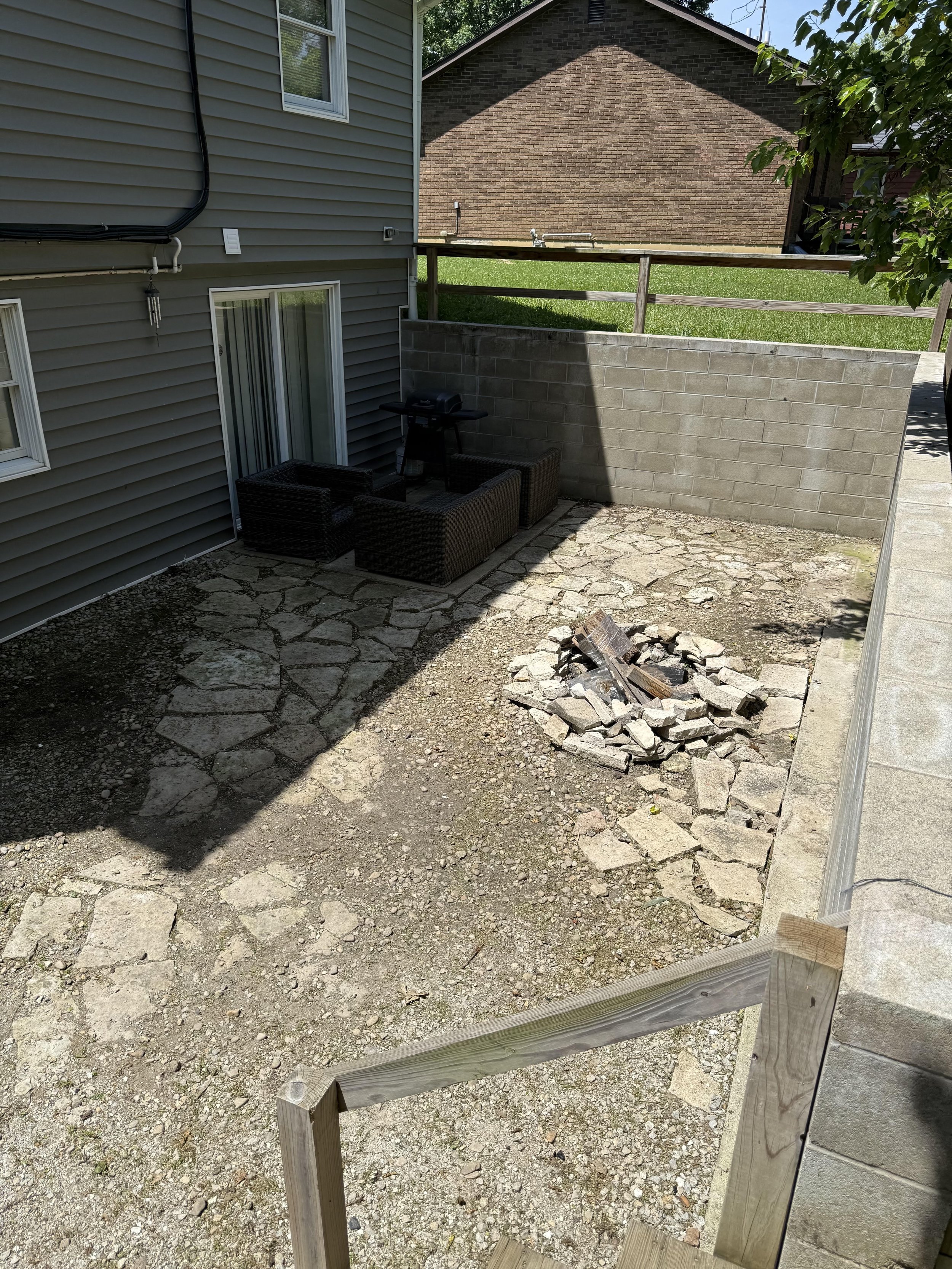 Backyard with outdoor seating area, small black grill, brick and vinyl house walls, gravel and stone ground, wooden privacy wall, and surrounding trees.