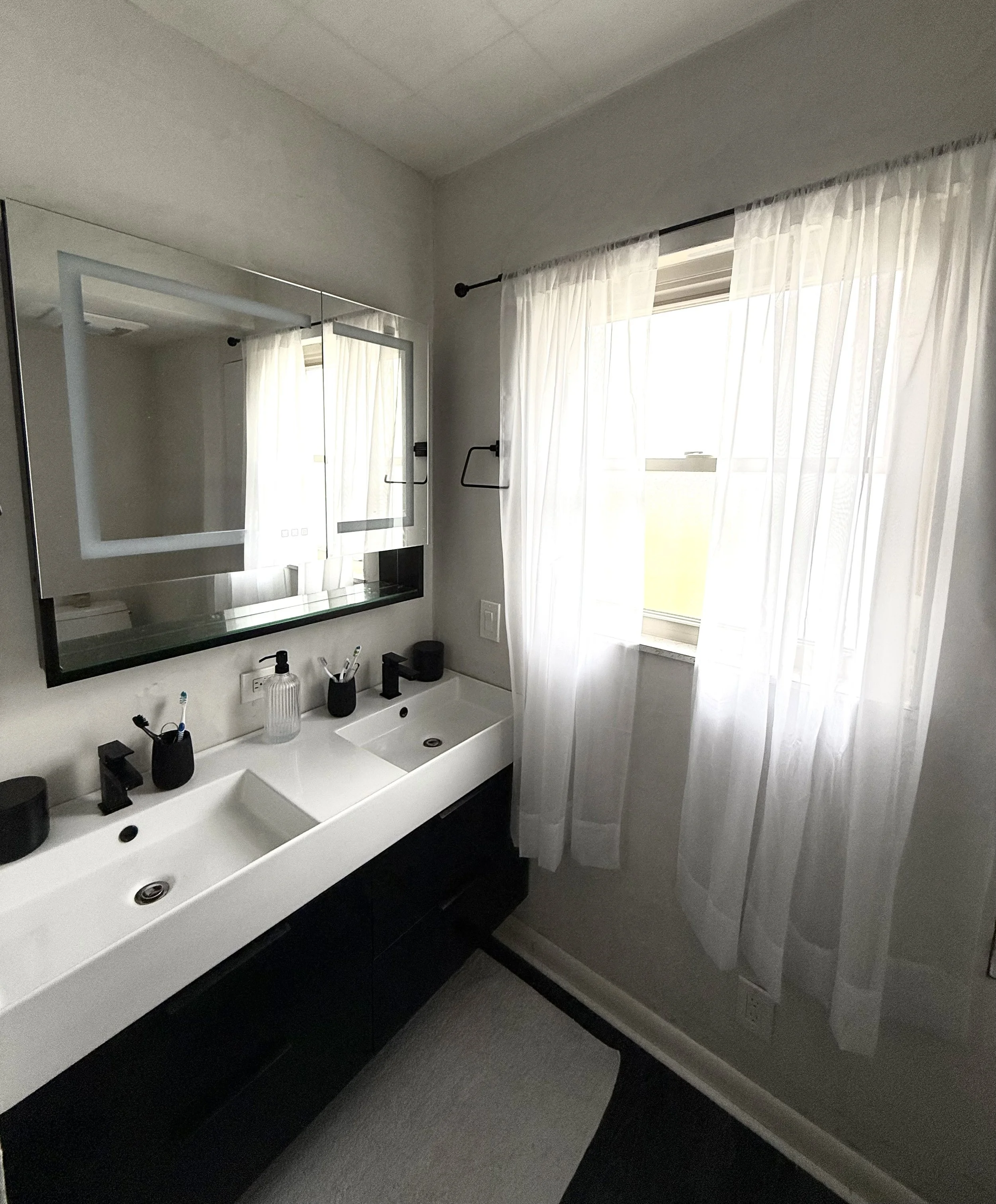 A modern bathroom vanity with a large mirror, white sink, black faucet, toothbrushes, and soap dispenser. The window has sheer white curtains allowing natural light to fill the space.