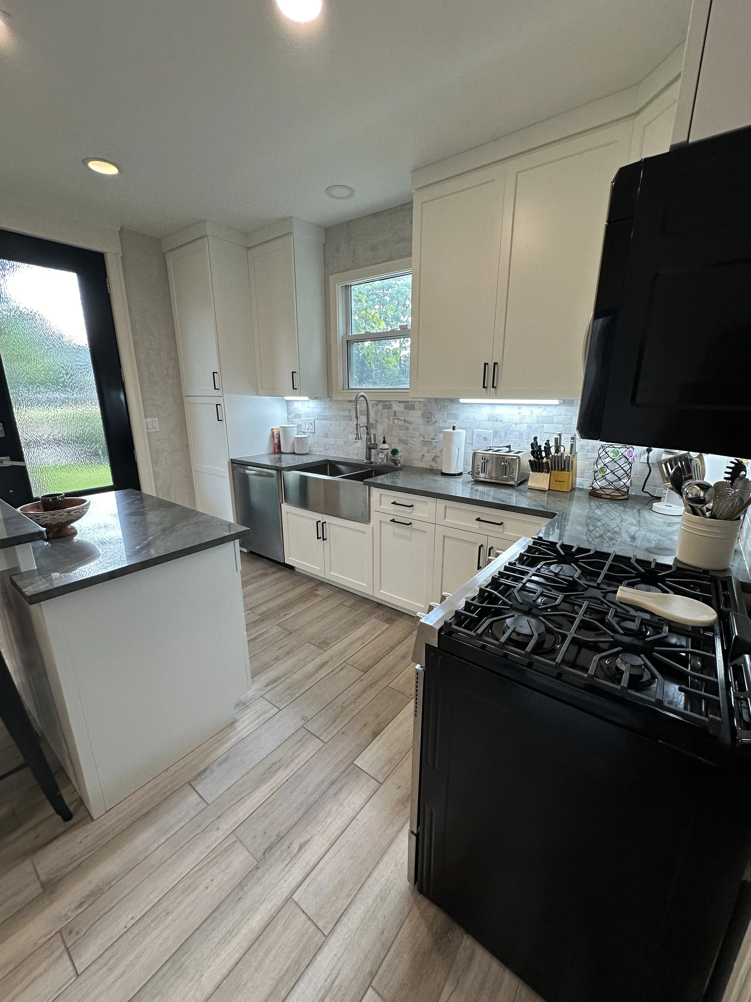 Modern kitchen with white cabinets, stainless steel sink, black appliances, and wood-look tile flooring, illuminated by natural and overhead lighting.