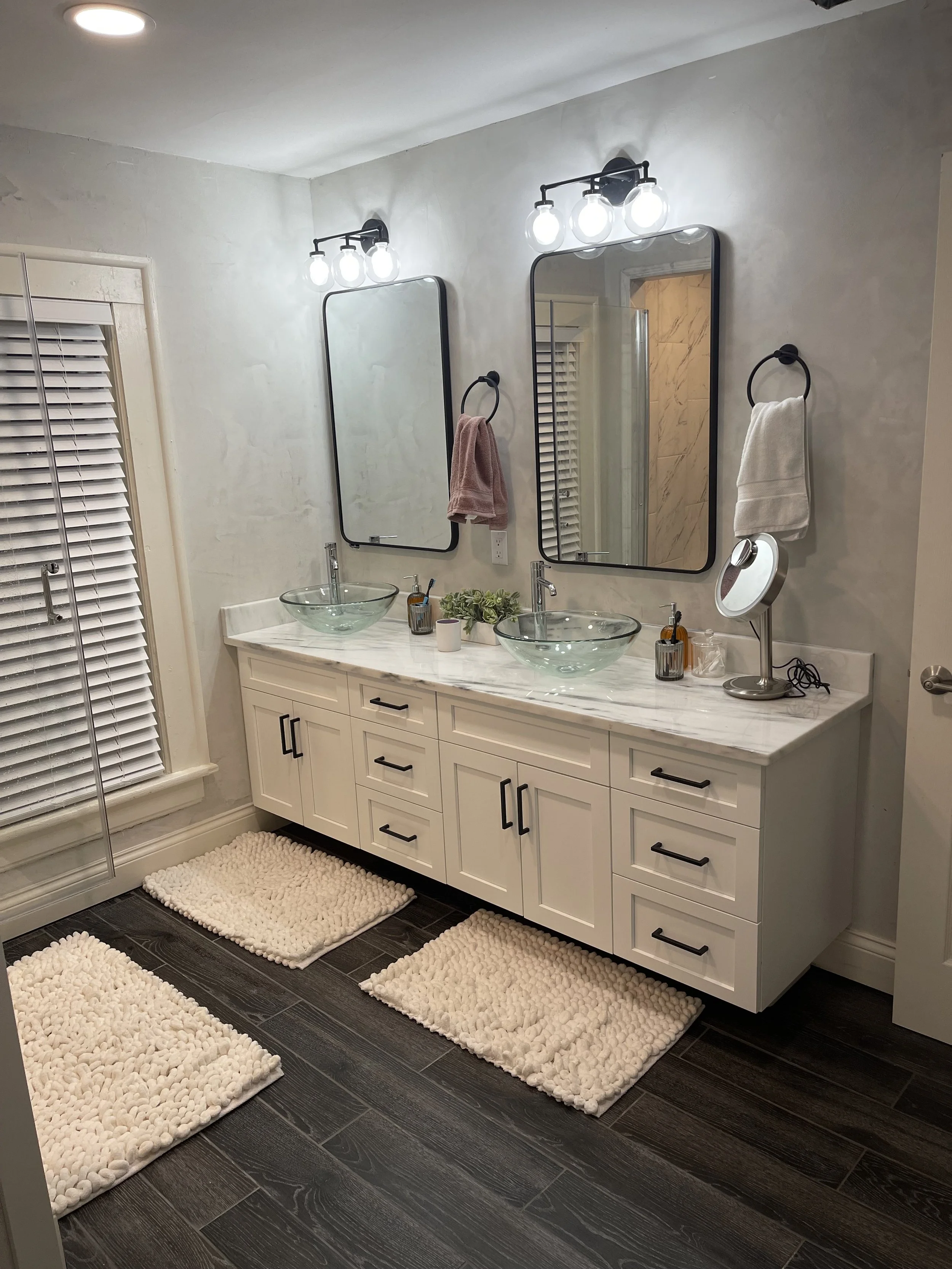 Modern bathroom with a white vanity, two glass vessel sinks, large mirrors, black fixtures, and plush cream rugs on dark wood flooring.