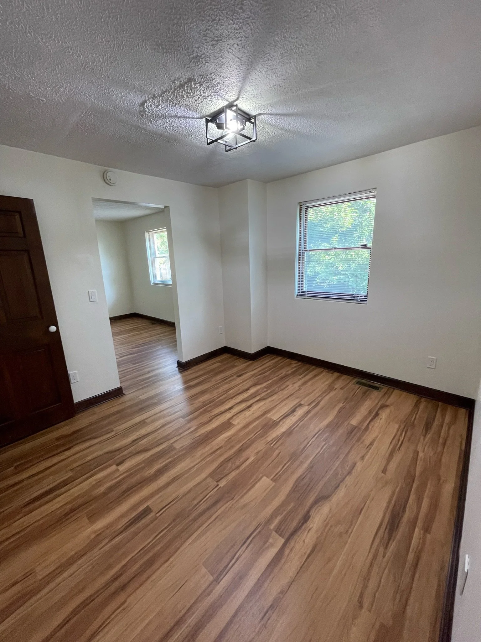 Empty room with wooden floors, beige walls, a window with blinds, and a ceiling light fixture.