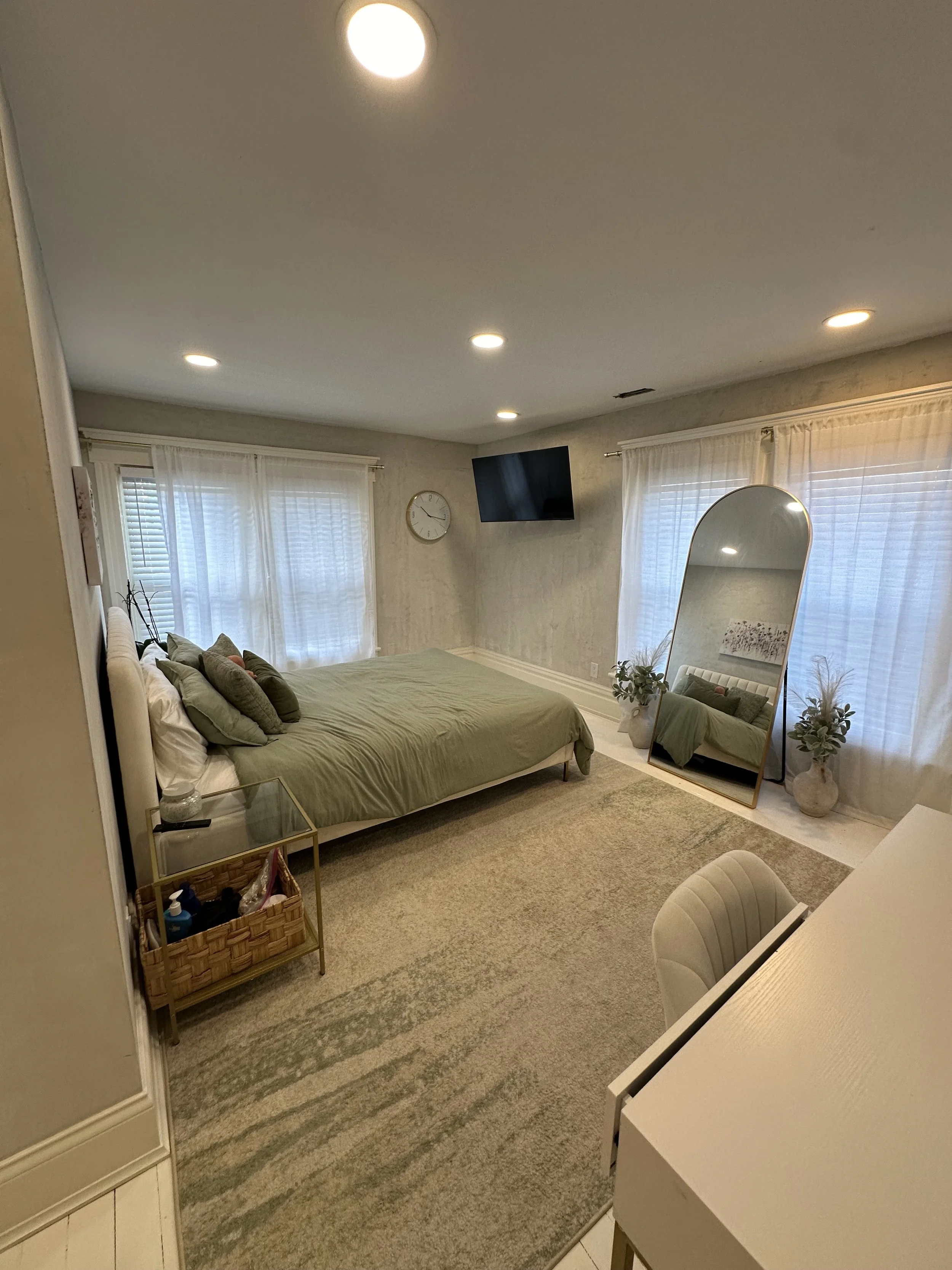 A cozy bedroom with a beige and green color scheme, featuring a bed with multiple pillows, a large mirror, a wall-mounted TV, a clock, potted plants, and windows with white curtains.