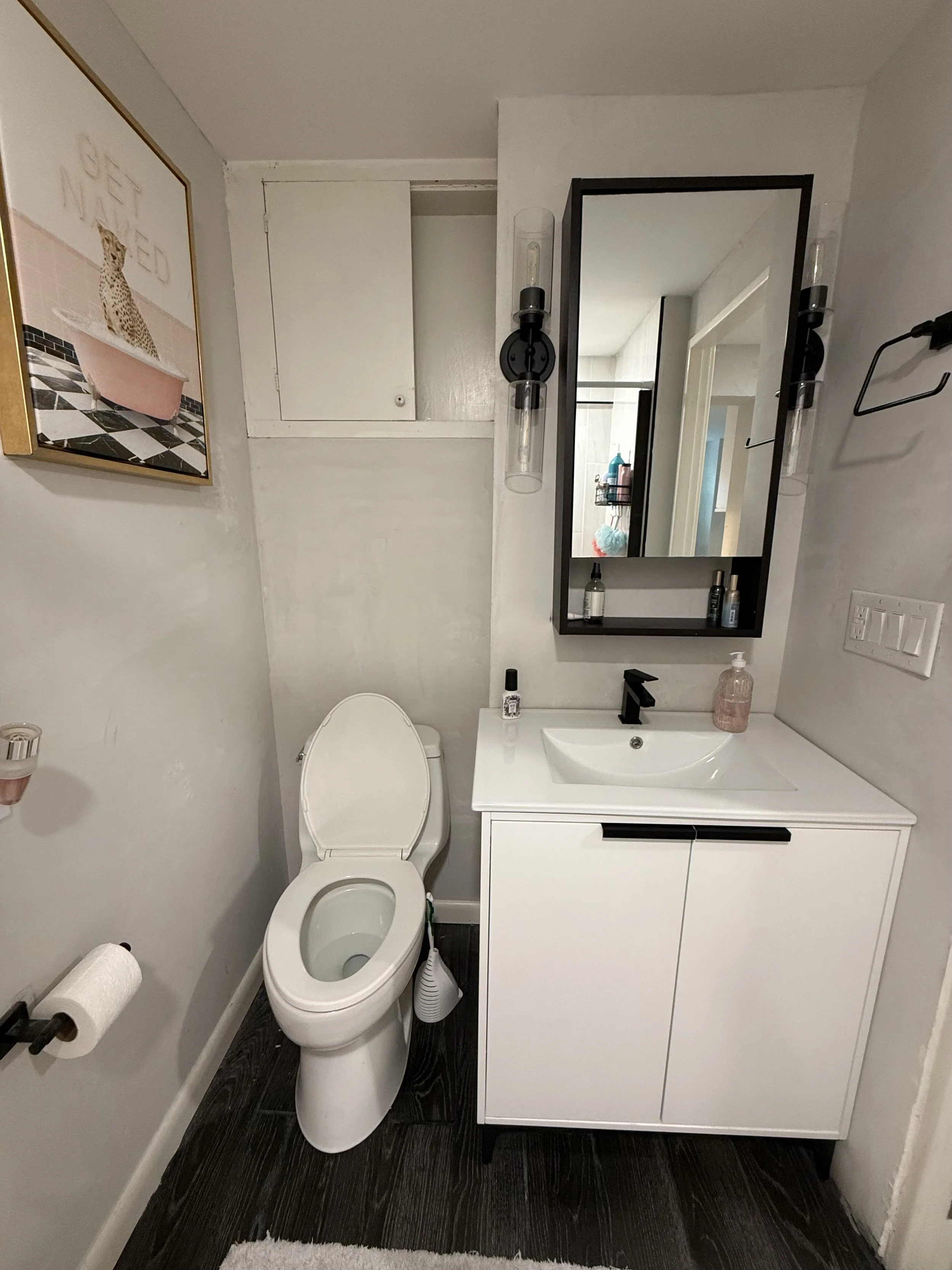 A small modern bathroom with a white toilet, white vanity with a black faucet, a mirror, and a dark wood floor. There is a framed picture of a cheetah on the wall and a towel rod with a toilet paper roll.