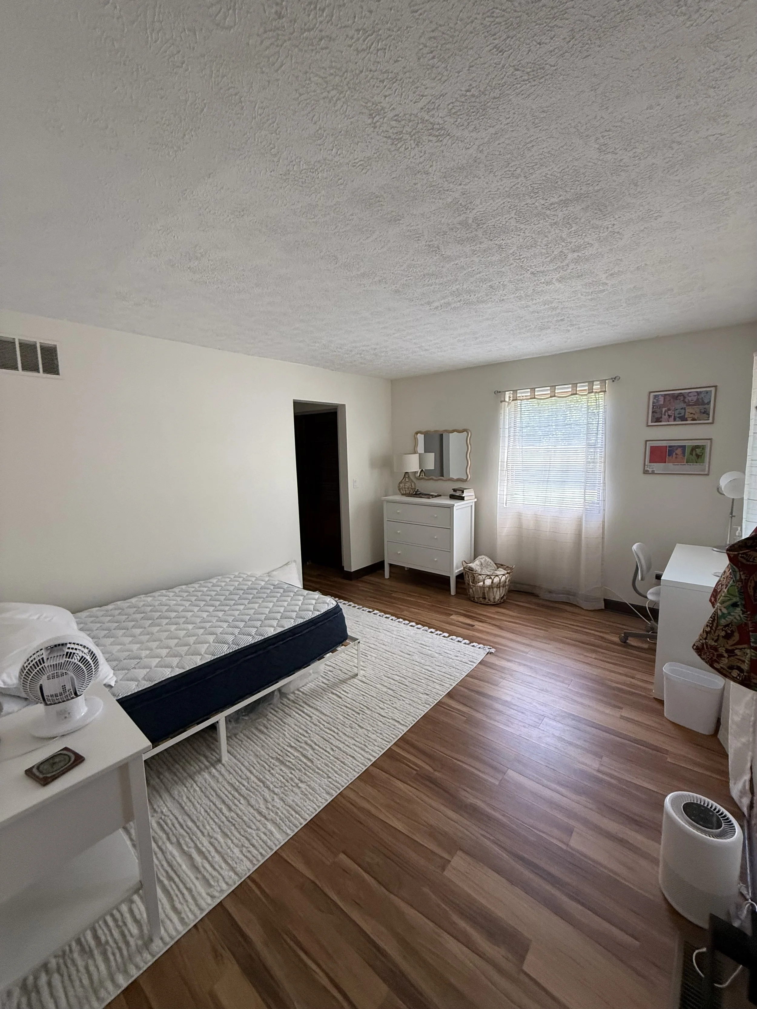 A bedroom with white walls, hardwood floors, a window with blinds, a white dresser, a framed mirror, artwork on the wall, and a bed with a mattress. A small table next to the bed has a fan on it, and there is a small wastebasket near the window.