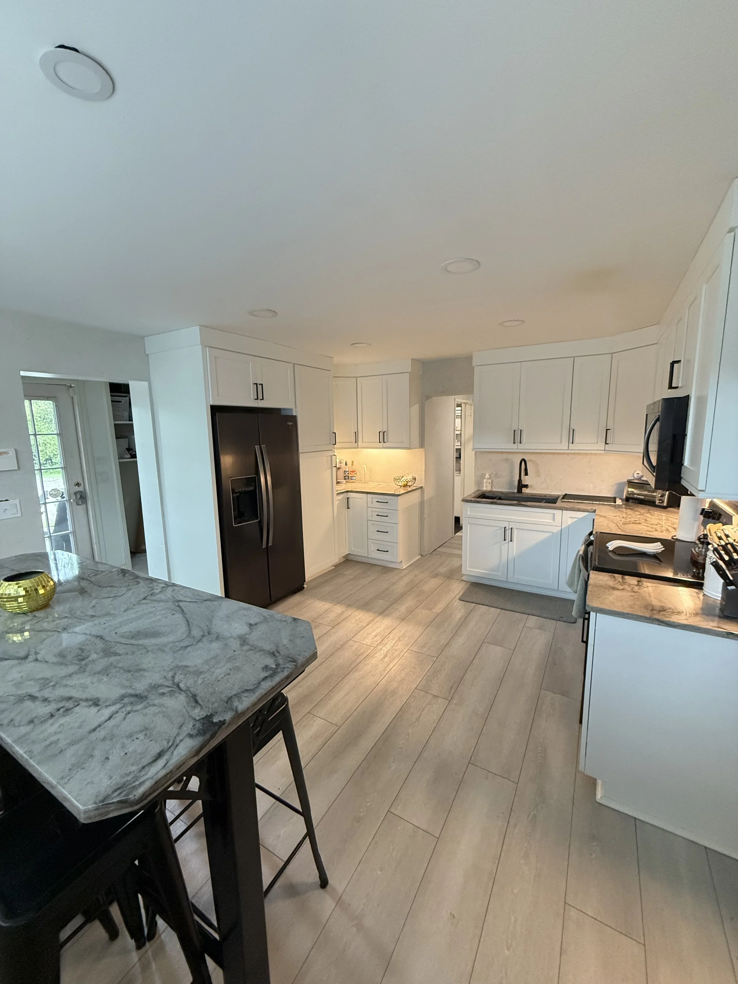 A modern kitchen with white cabinets, stainless steel refrigerator, granite countertops, and a tiled floor. Part of a marble kitchen island with two black chairs is visible in the foreground.