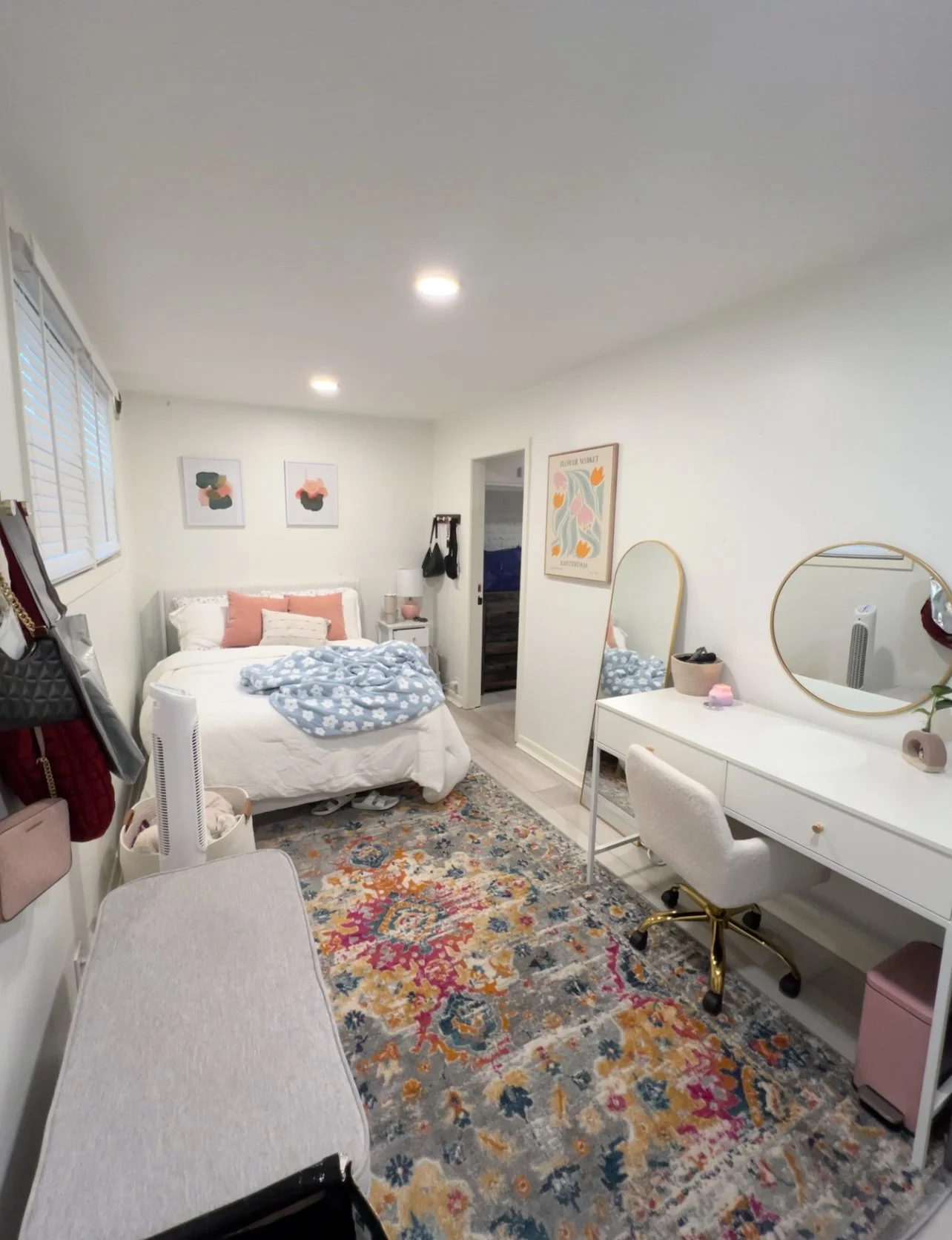 A cozy bedroom with a bed, white bedding, pink pillows, and a blue patterned blanket. There are two wall art pieces above the bed, a white side table to the right of the bed, and a small mirror on the wall. A colorful rug covers part of the floor. On