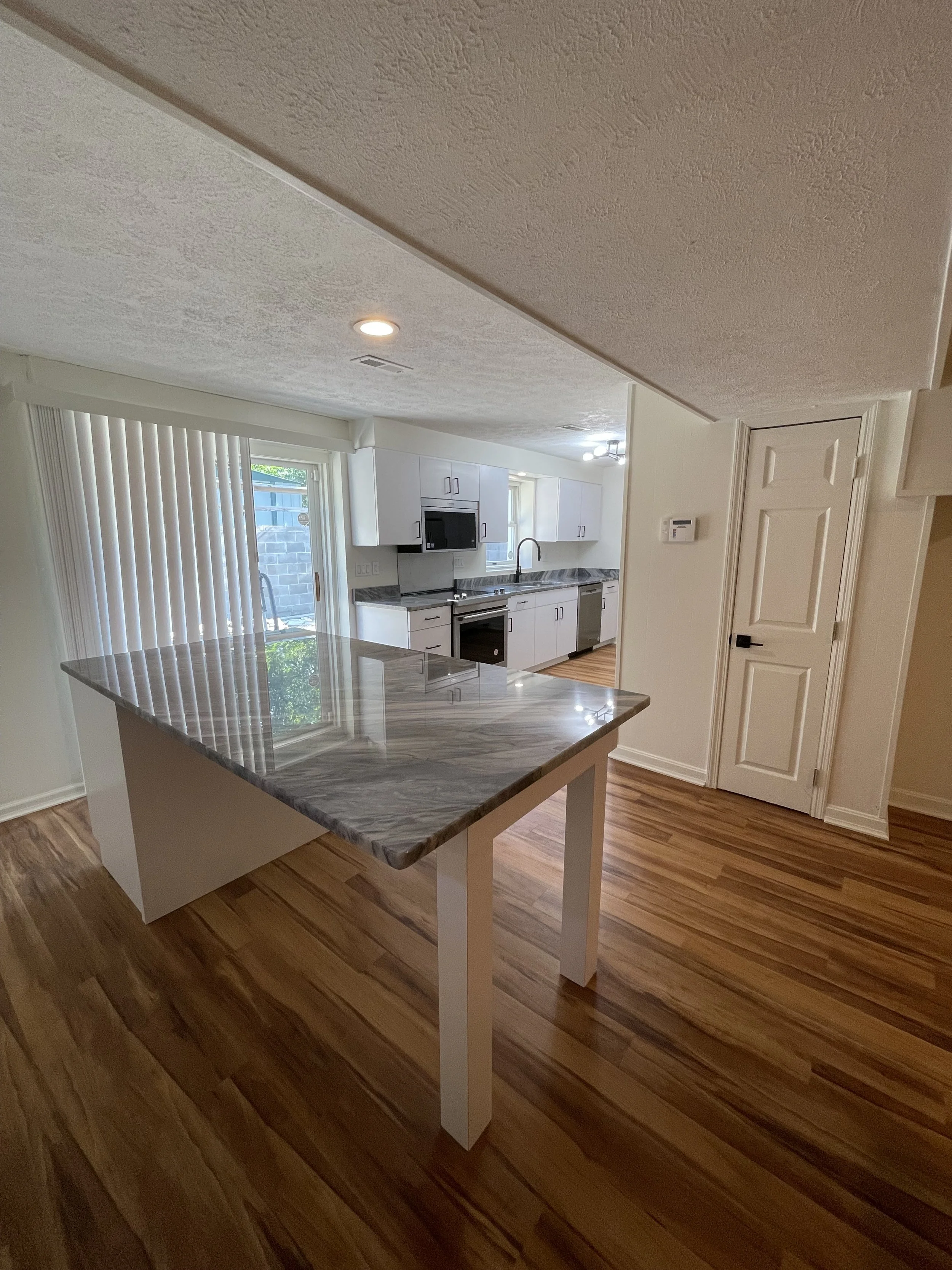 A kitchen with white cabinets, a marble countertop on a kitchen island, hardwood floors, a sliding glass door with vertical blinds, and a ceiling with recessed lighting and a ceiling fan.