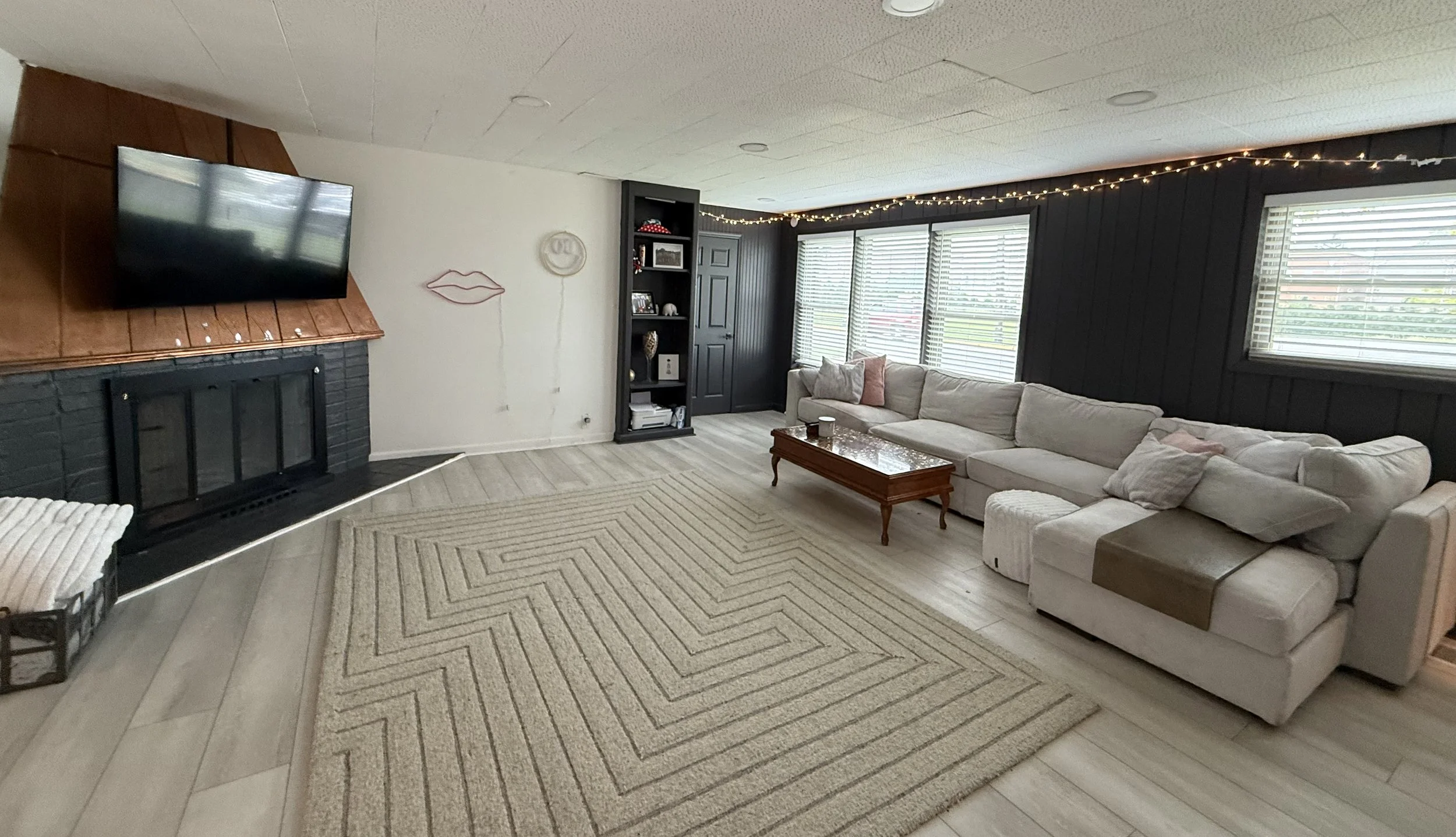Living room with a beige sectional sofa, a wooden coffee table, a large window with blinds, a black built-in bookshelf, a fireplace with a copper-colored canopy, and string lights on the black wall.