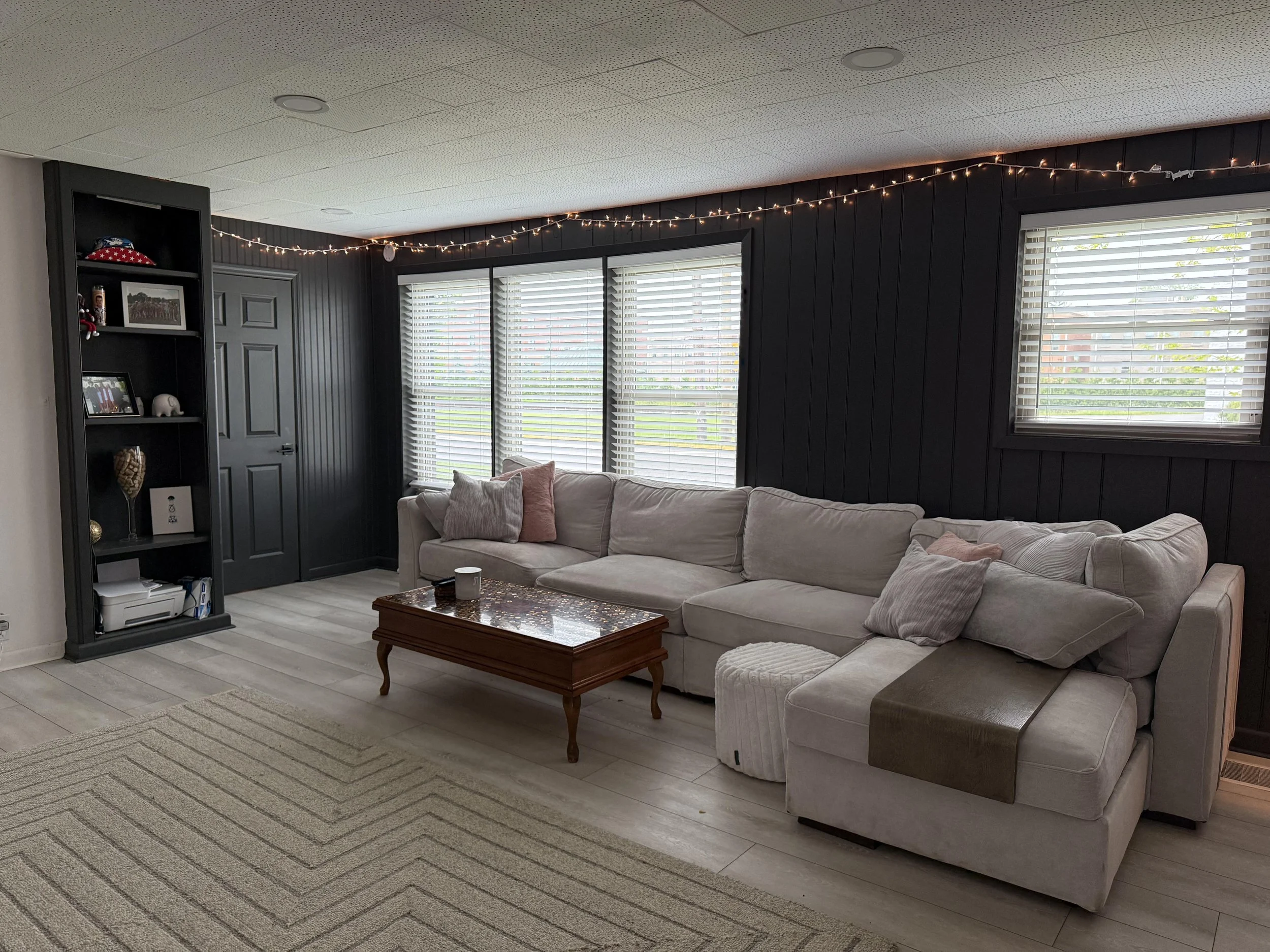 Living room with black paneled walls, three large windows with blinds, a beige sofa with pink cushions, a wooden coffee table, a gray armchair, a black bookshelf with decorative items, and fairy lights hanging along the wall.