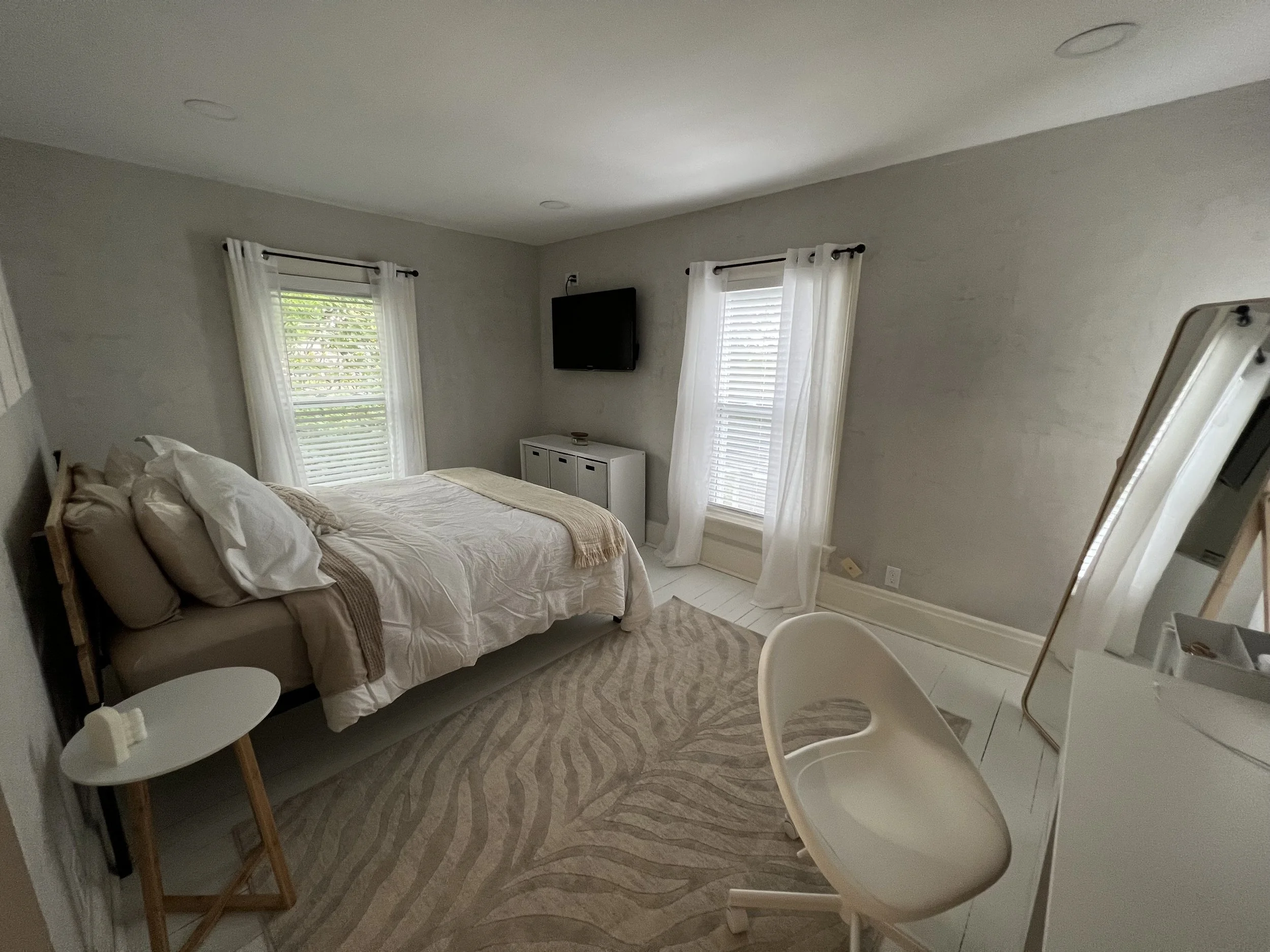 A bedroom with a bed, two windows with white curtains, a wall-mounted TV, a white cabinet, a white chair, a round side table, and a patterned rug.