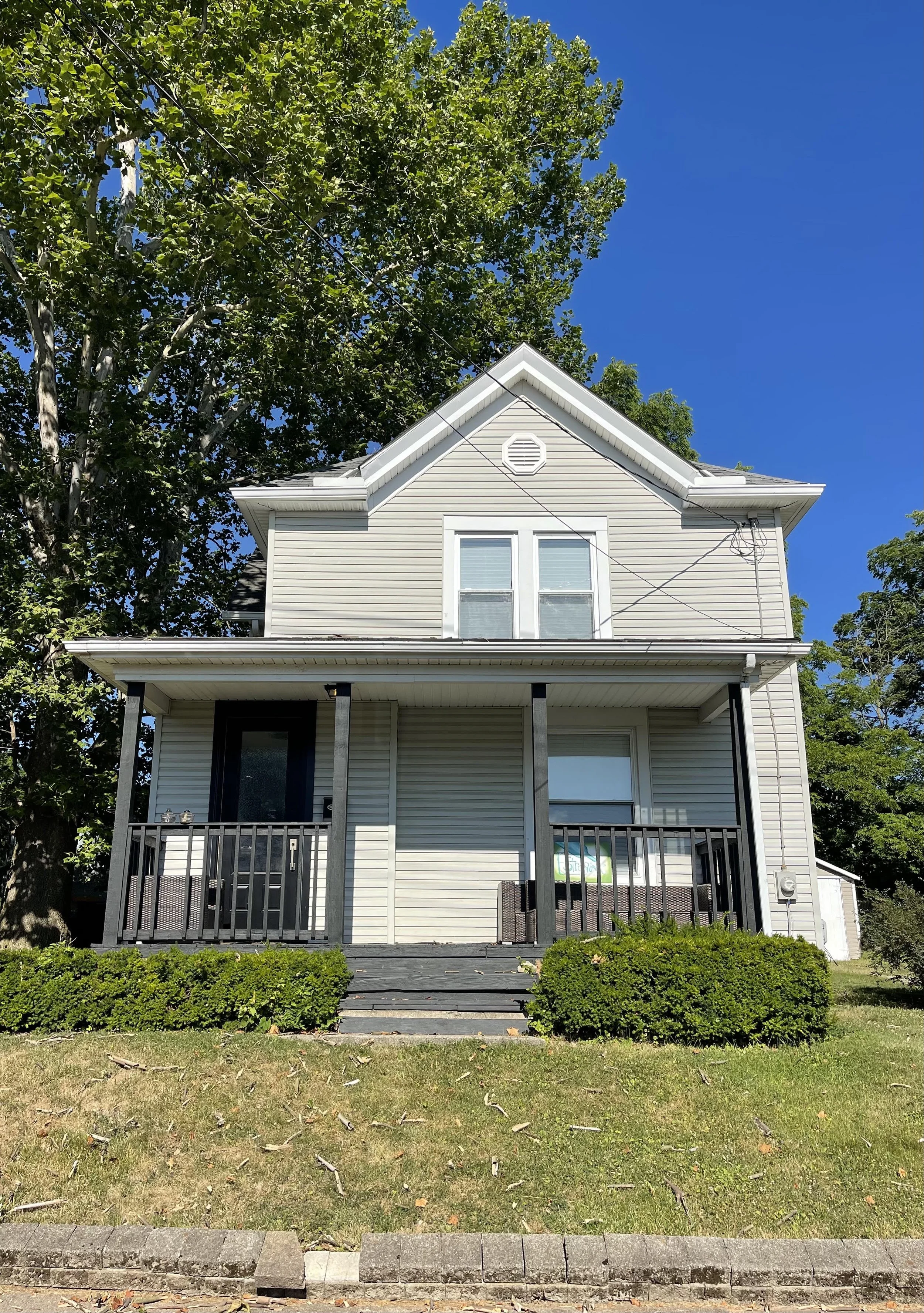 A two-story house with light-colored siding, a porch with black railing, and stairs leading to the entrance, set against a clear blue sky. There are green shrubs and a tree in the front yard.
