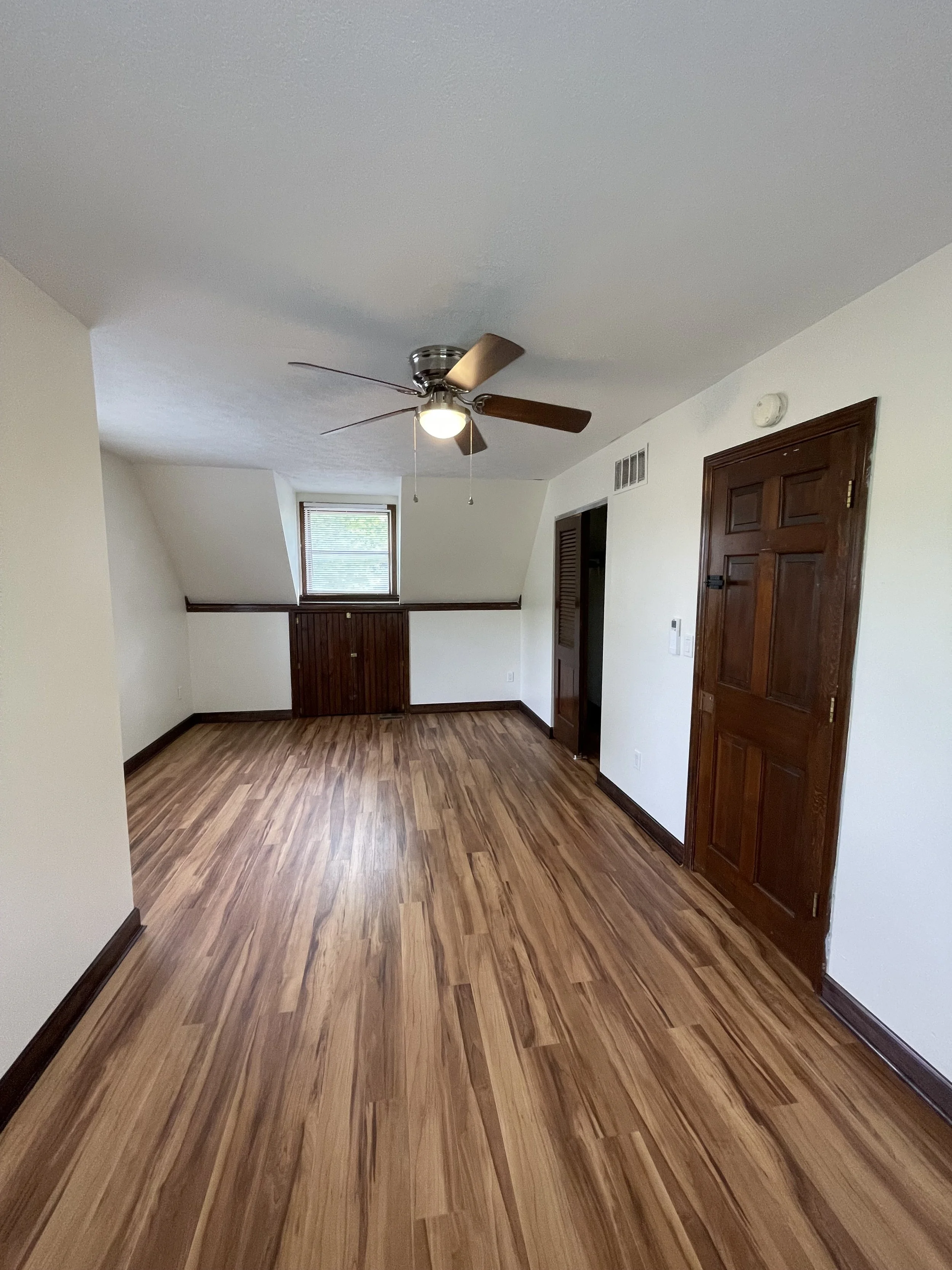 A room with wood flooring, a ceiling fan with light, a small window with blinds, white walls, and brown wooden doors and trim.
