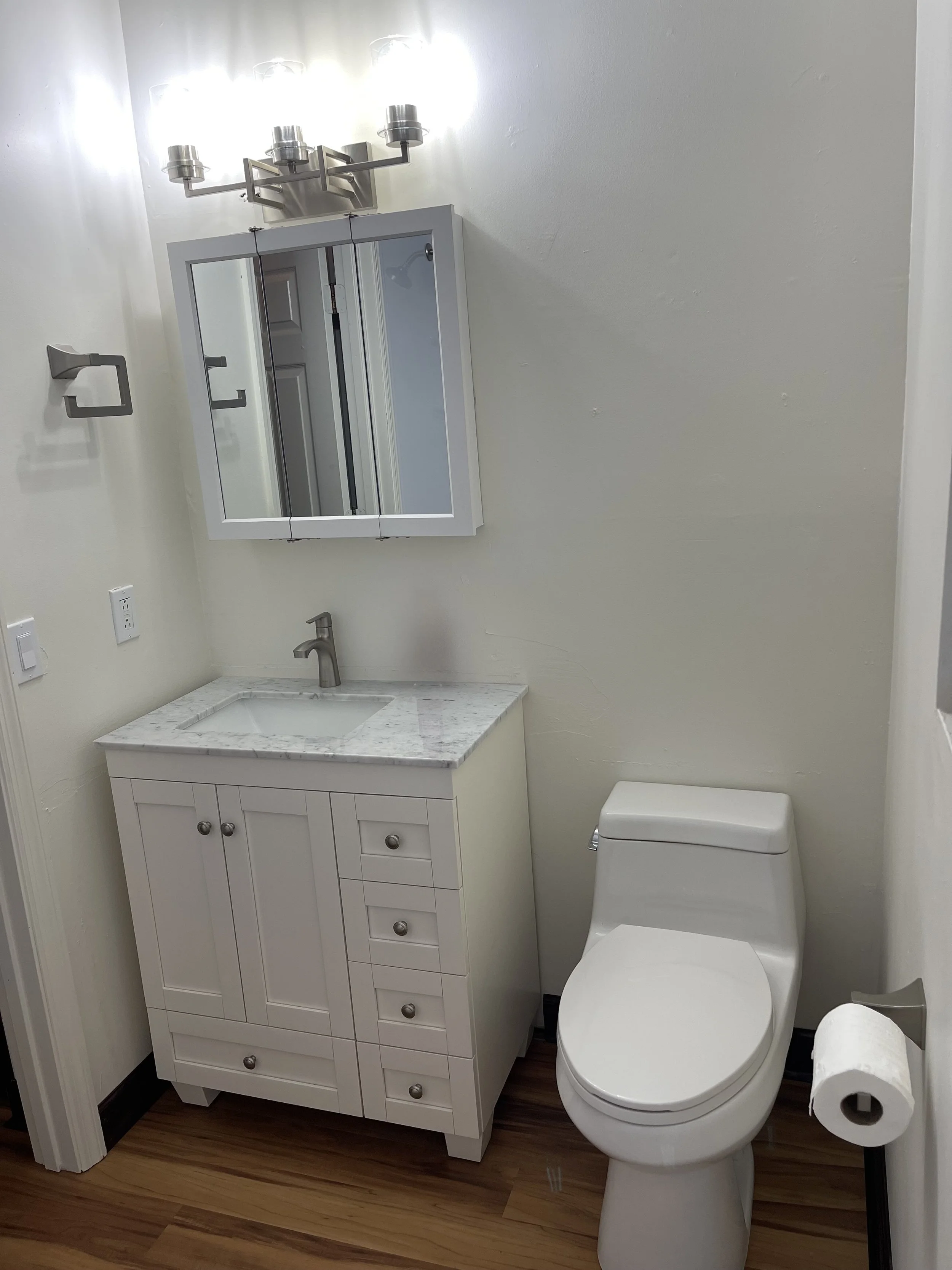A small bathroom with a white vanity with drawers and a marble countertop, a silver faucet, a wall-mounted mirror cabinet, a modern silver toilet paper holder with a roll of toilet paper, a toilet, and a roll of toilet paper on a holder. The walls ar