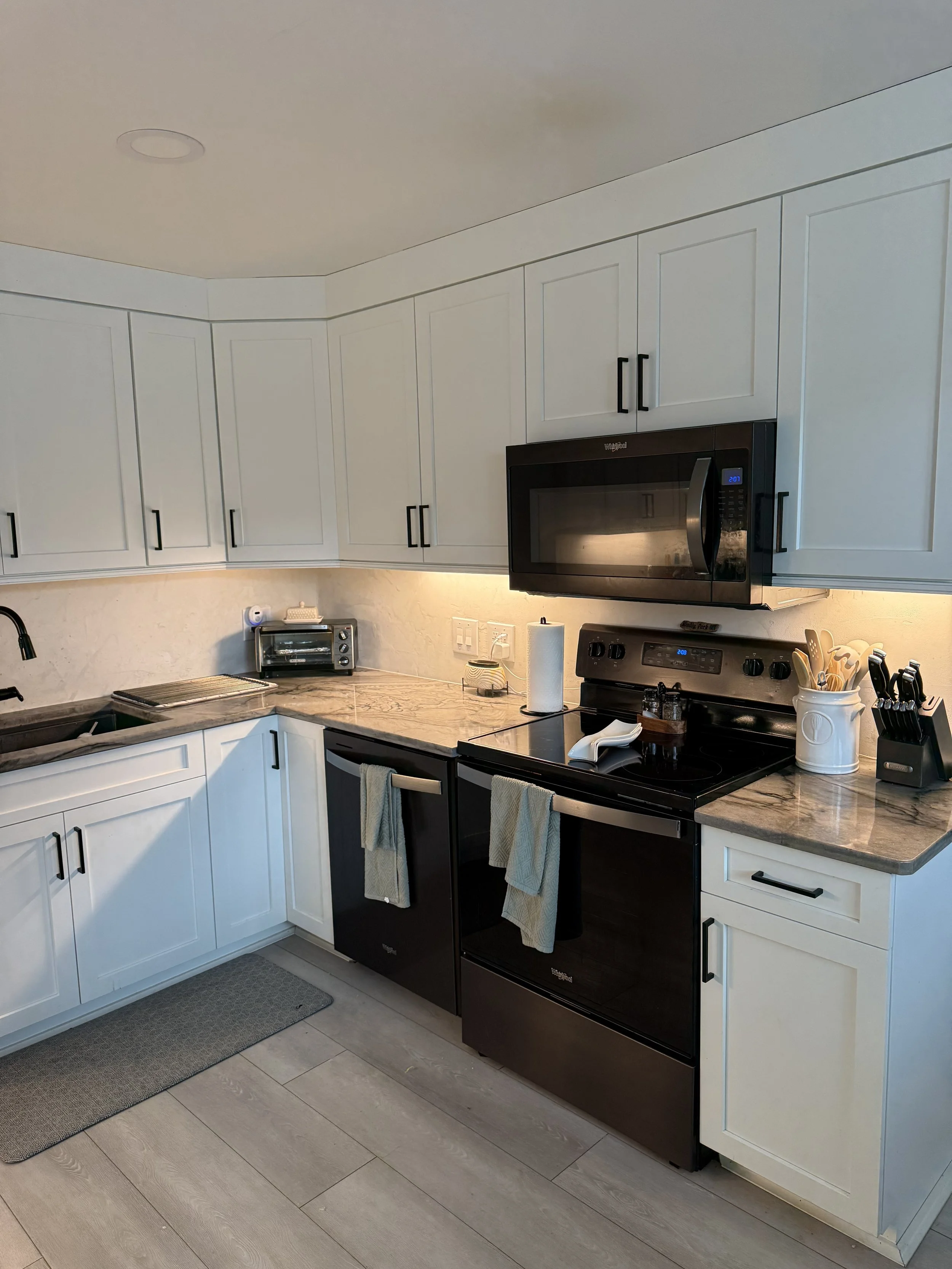 A modern kitchen with white upper cabinets, black lower cabinets, a black microwave, and a marble countertop. There are kitchen tools, paper towels, and small appliances on the counter.