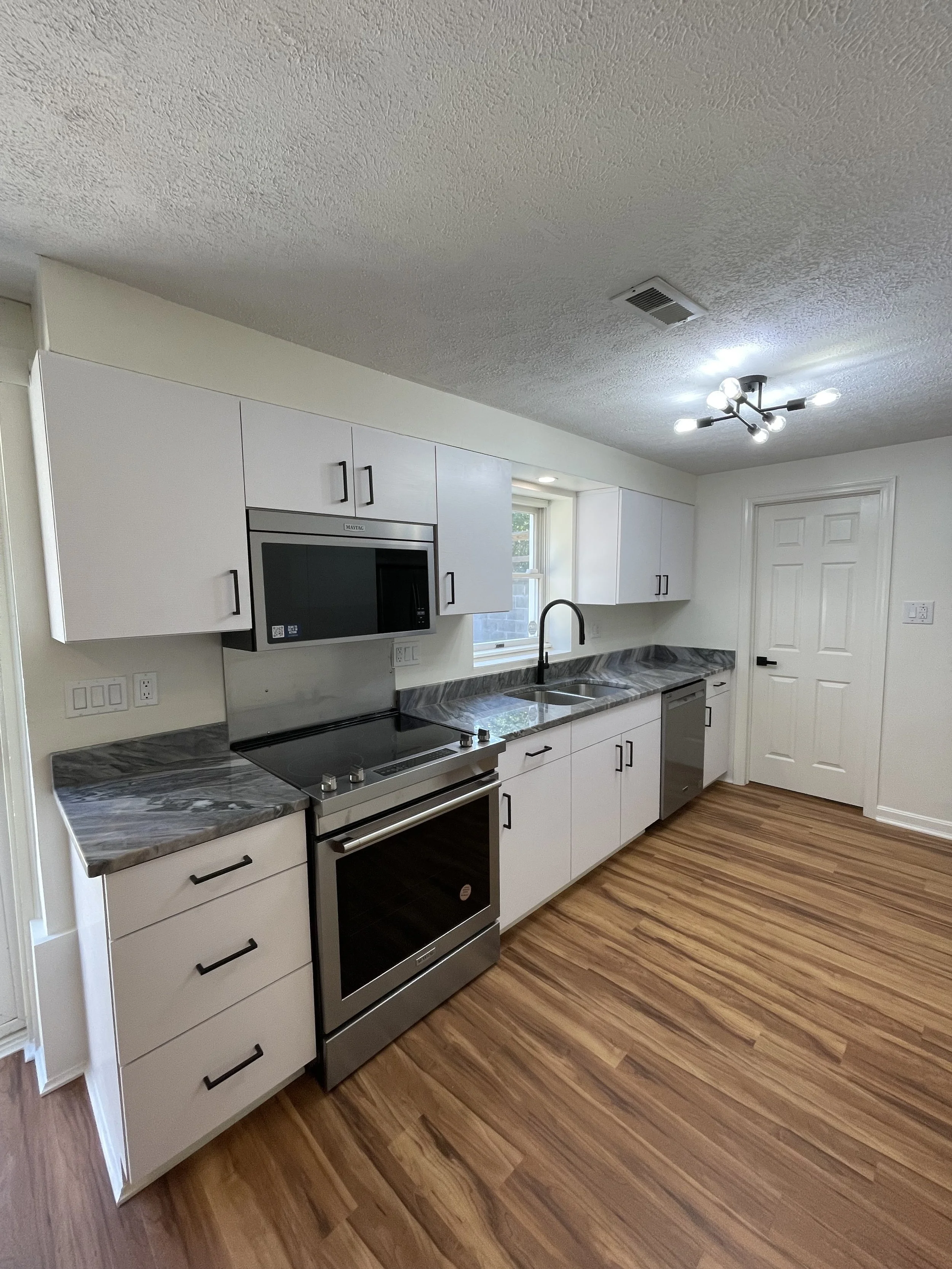 A modern kitchen with white cabinets, a gray countertop, a black microwave, a stainless steel oven, a black stove, and wood laminate flooring. There is a window above the sink and a white door to the side.