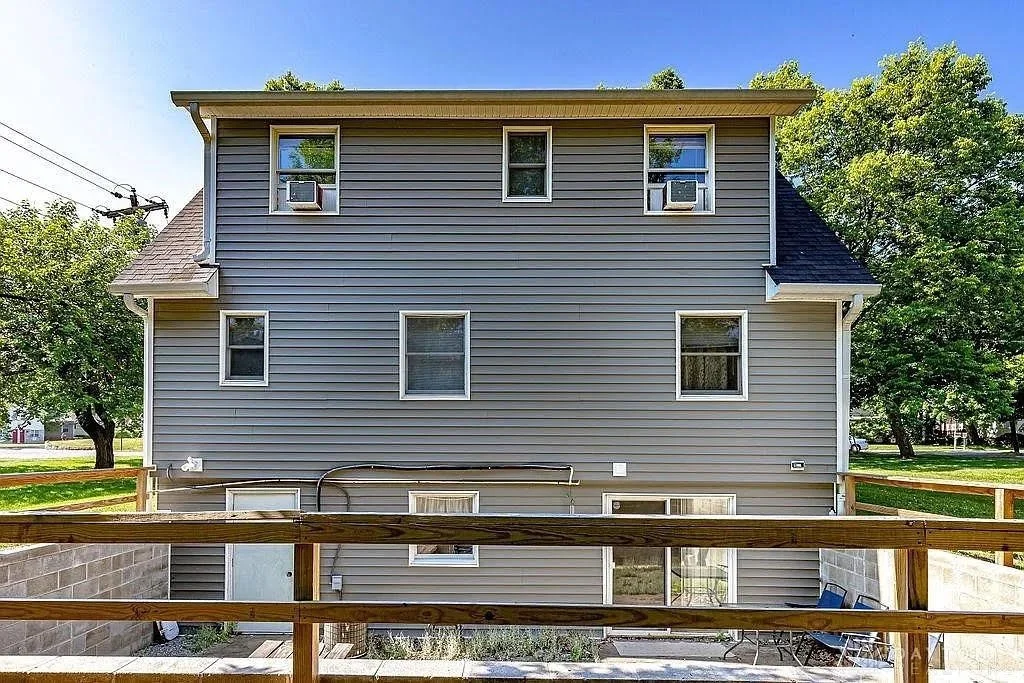 Front of a three-story gray house with multiple windows and air conditioning units, surrounded by green trees and a wooden fence in the foreground.