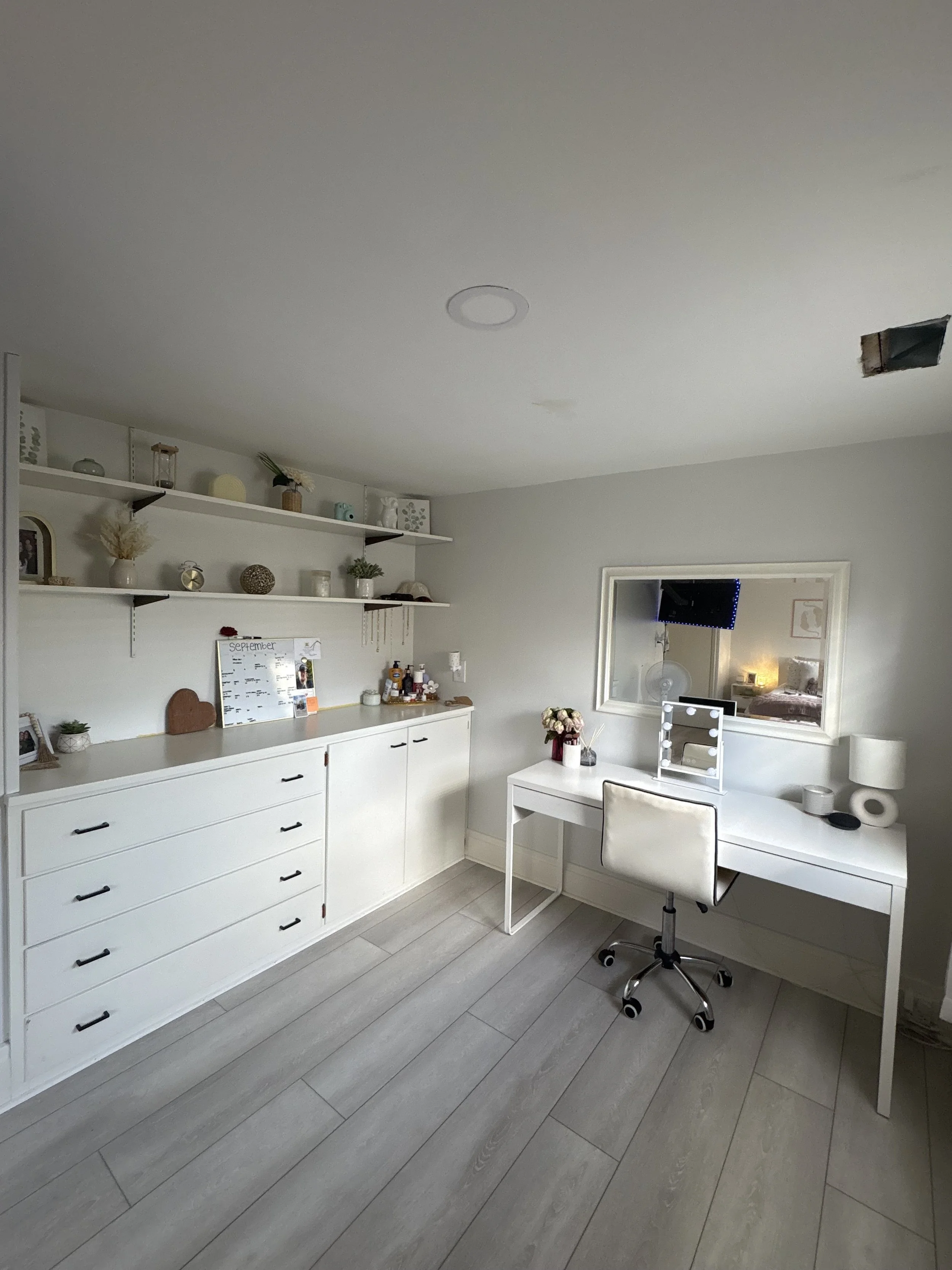 A bright, minimalist home office with a white desk and chair, wall-mounted white shelves decorated with plants and ornaments, a mirror reflecting a bedroom, and light gray wooden flooring.