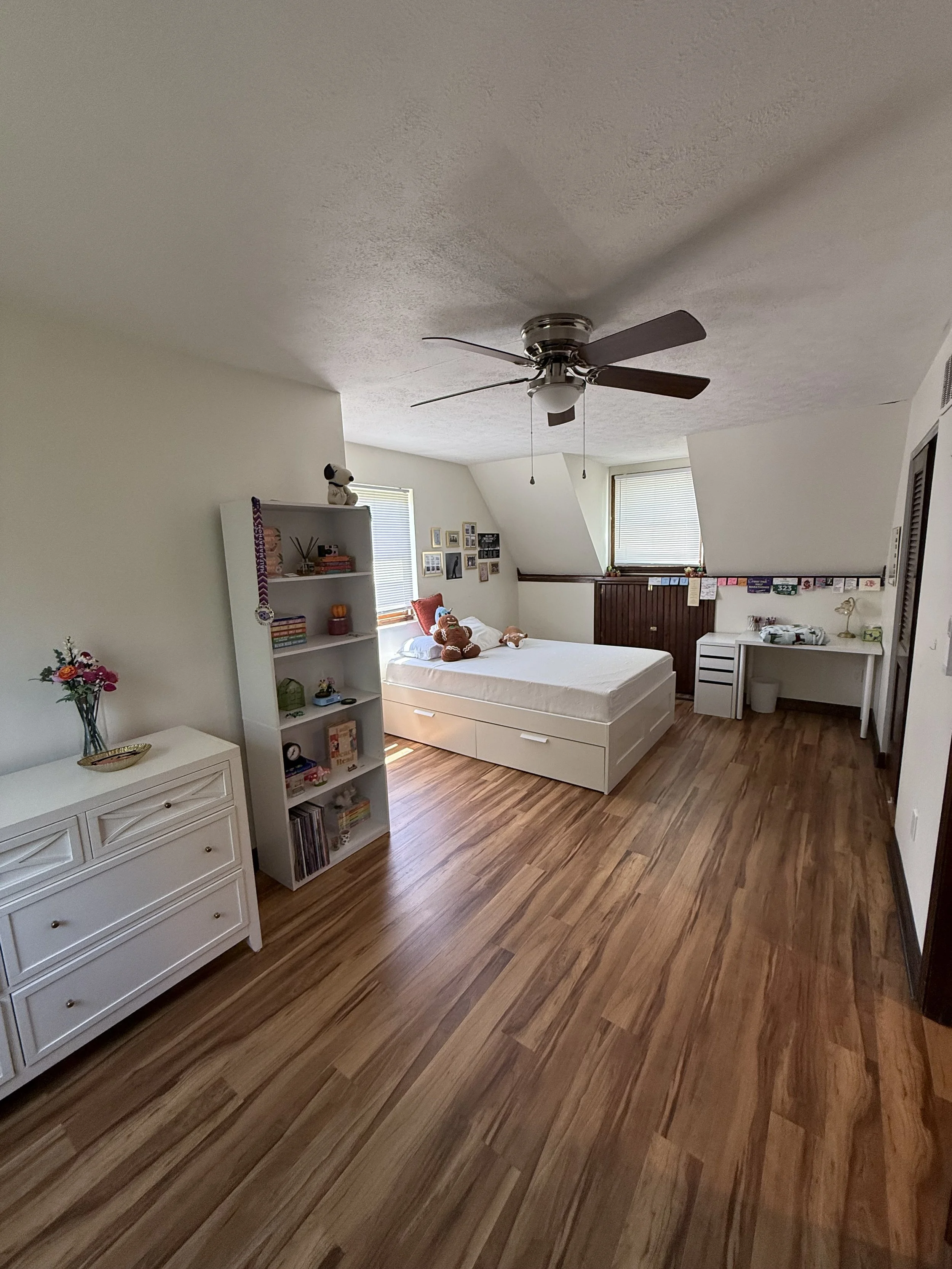 A bedroom with a white bed, a ceiling fan with lights, two windows with blinds, a white dresser with a vase of flowers, and a white bookshelf with toys and books. The floor is wood, and the walls are painted light.