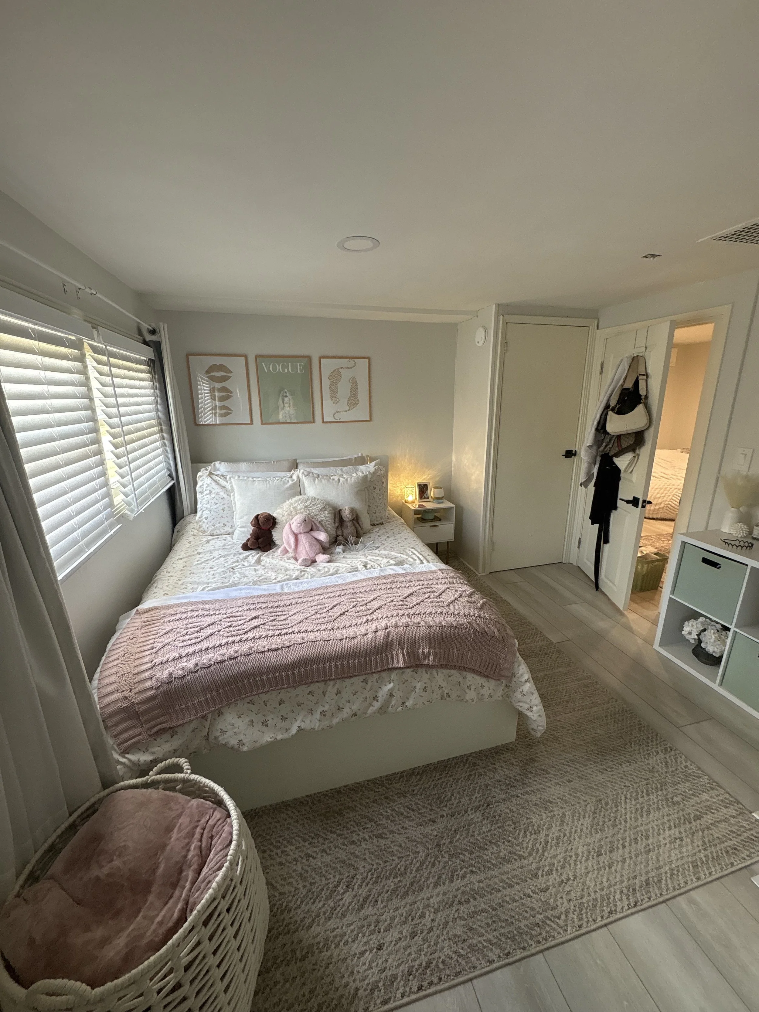 A cozy bedroom with a bed adorned with plush toys and a pink throw blanket, wicker basket at the foot of the bed, framed wall art, window with blinds, small nightstand with lamp, and an open door leading to a bathroom.