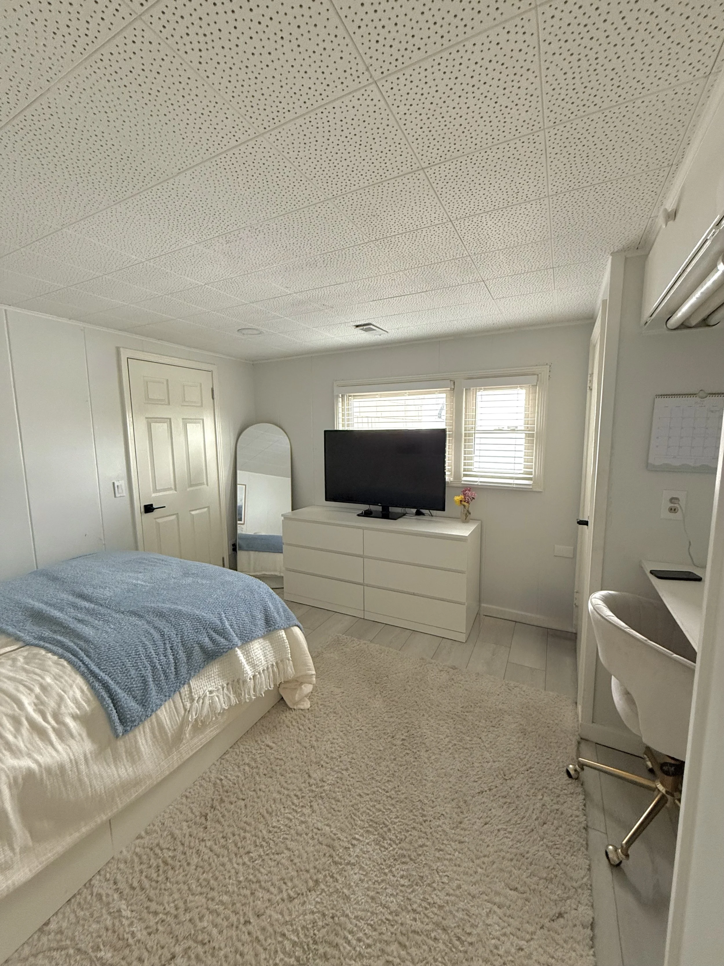 A bedroom with a bed covered with a light-colored bedspread and a blue blanket, a beige carpet, a white dresser with a TV on top, a small mirror, two windows with blinds, and a white wall-mounted calendar.