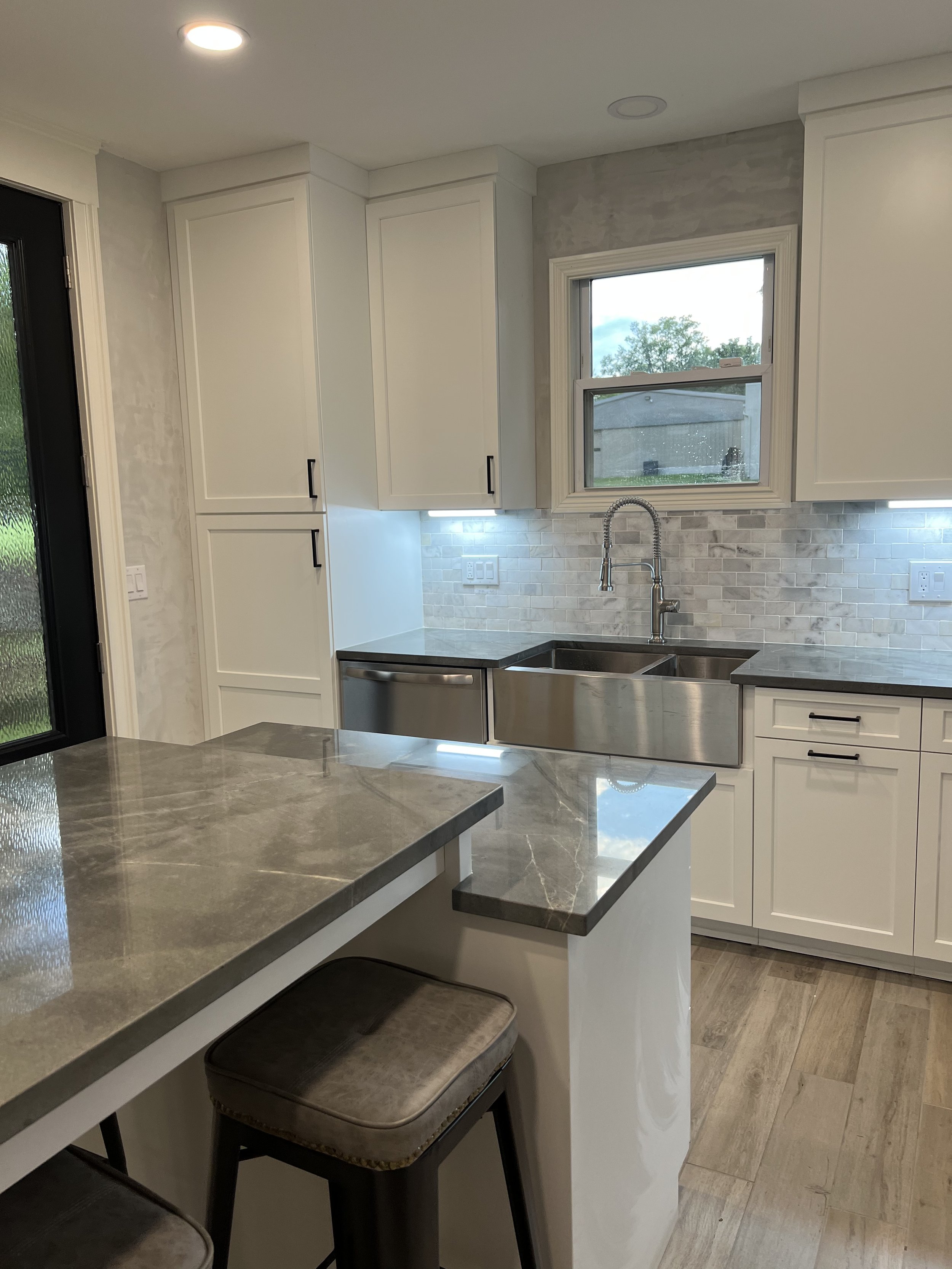 Modern kitchen with white cabinets, a double sink with a chrome faucet, a gray marble countertop island, a window above the sink, a brick backsplash, and wood-look flooring.